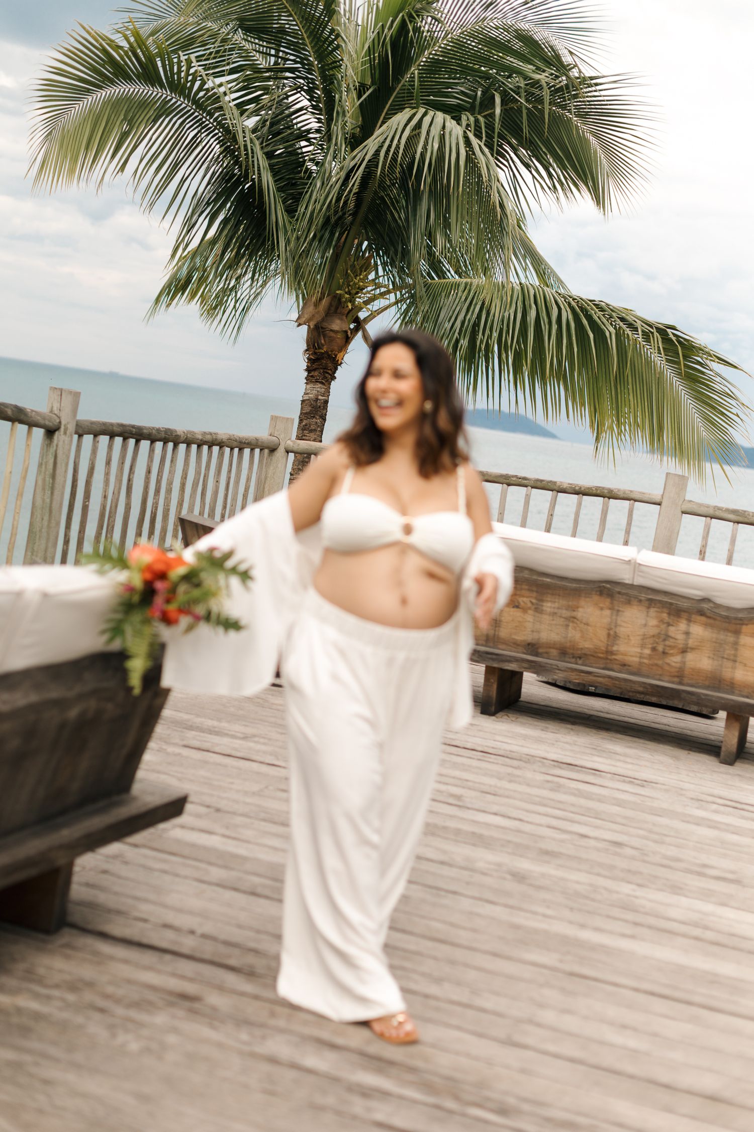 A person in a white dress twirls on a wooden deck with palm trees and ocean views in the background.