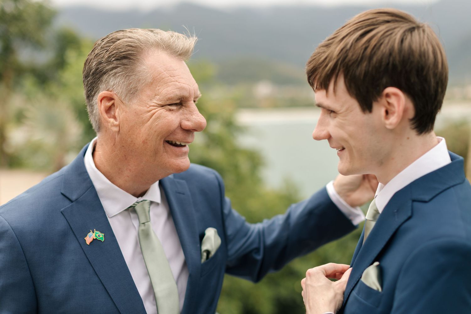 Two people in formal attire share a candid moment of laughter outdoors with mountains in the background.