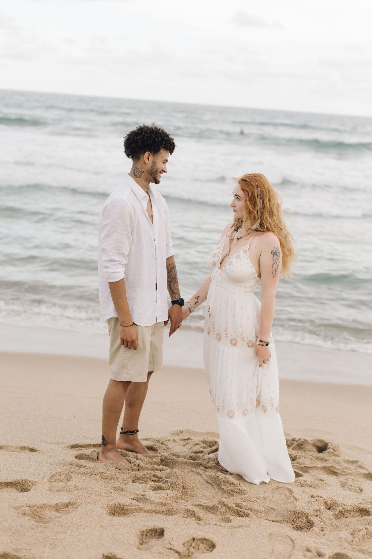 A couple in white outfits walks together on a sandy beach with waves in the background.