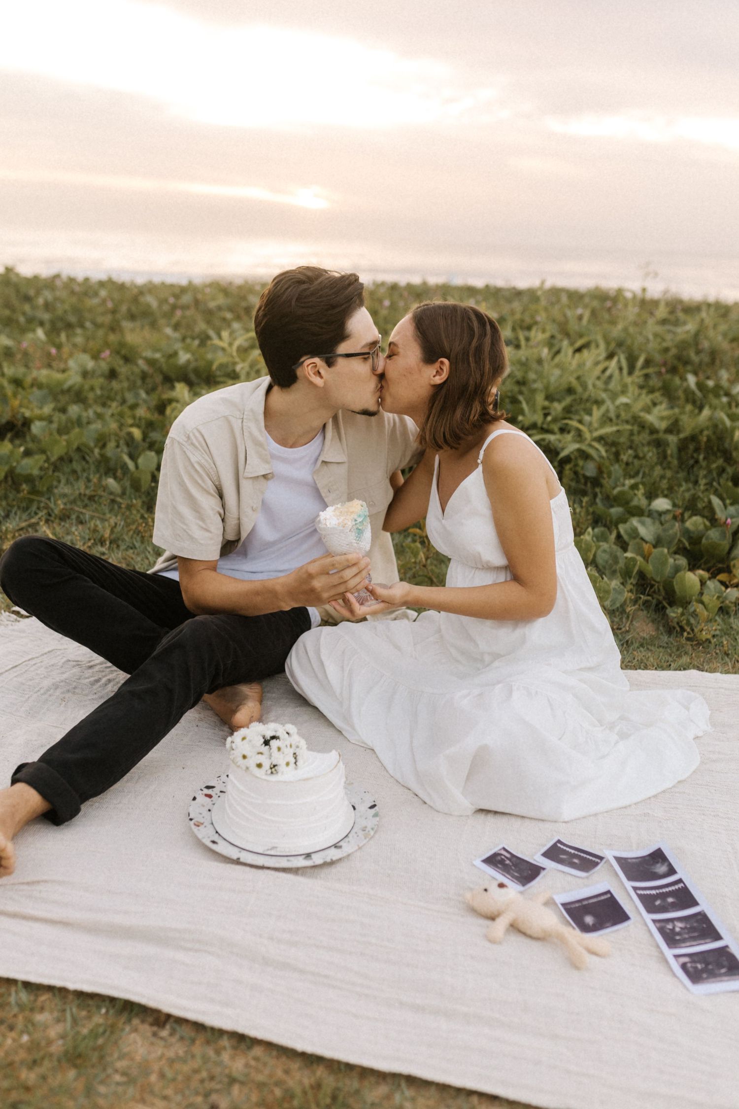 A romantic couple shares sweet moments on a picnic blanket with cake and photos at sunset.
