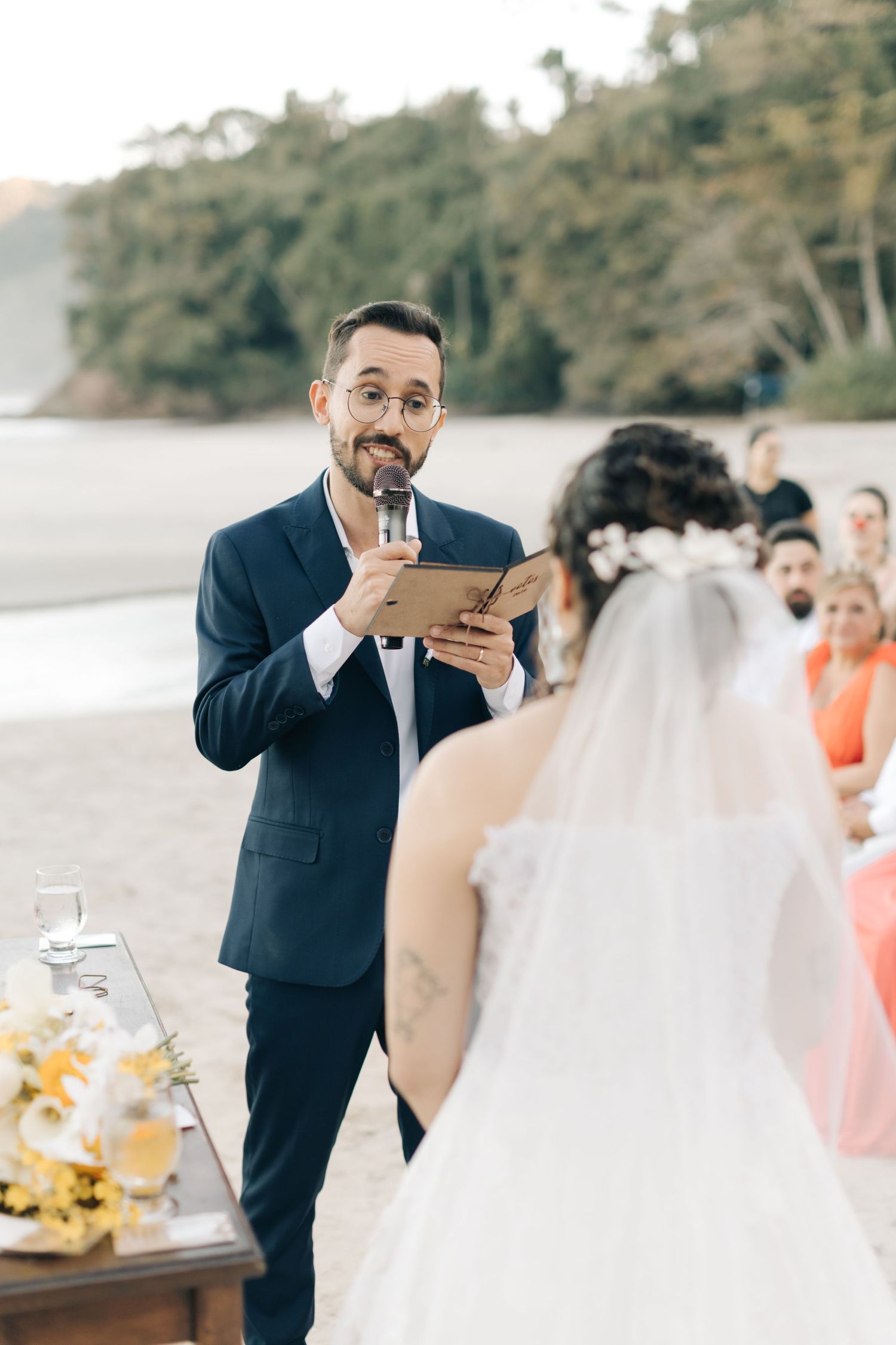 Officiant in navy suit delivers ceremony speech at beachside wedding.