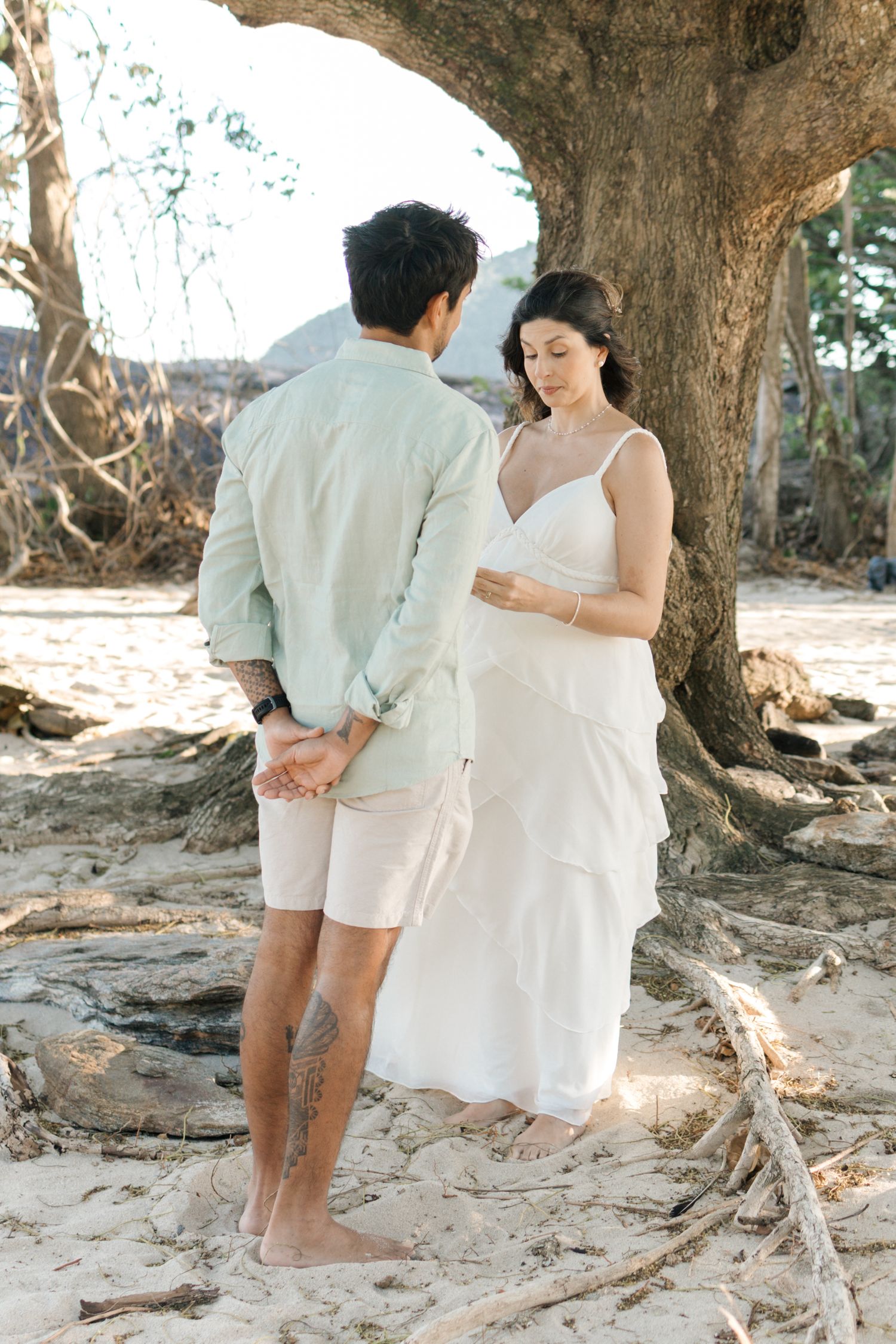 A couple in white clothing embraces on a rocky beach beneath a large tree during a romantic photoshoot.