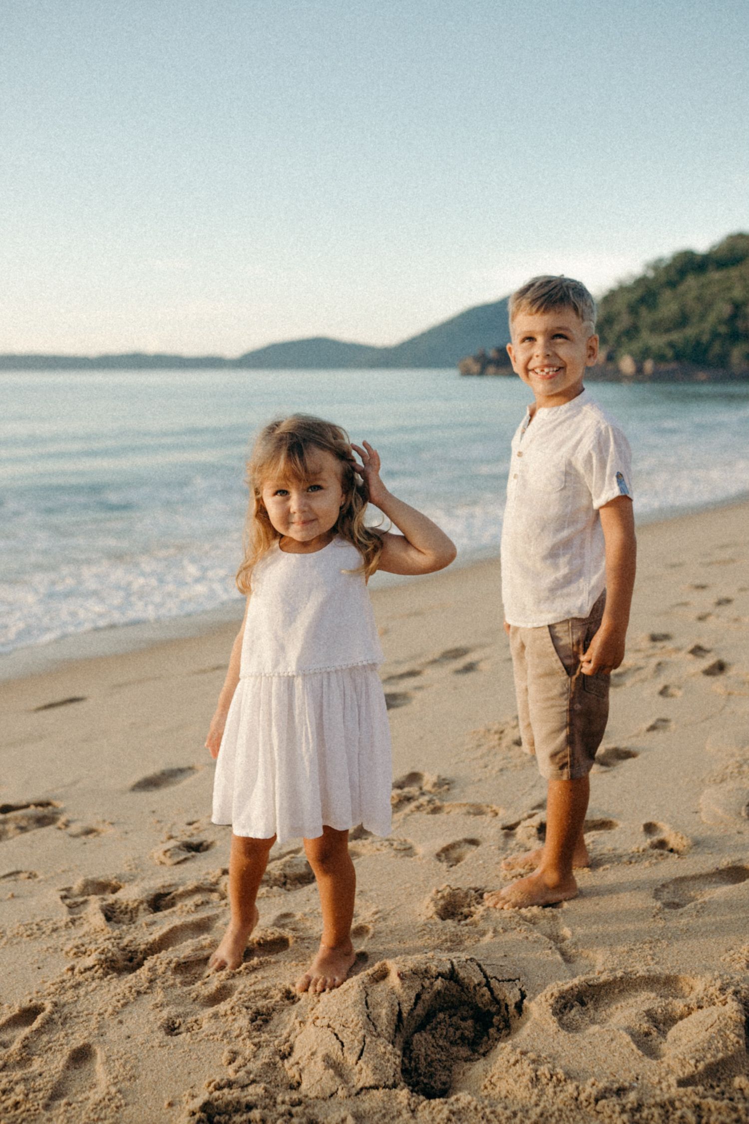 Two children in white outfits standing on beach with mountains in background.
