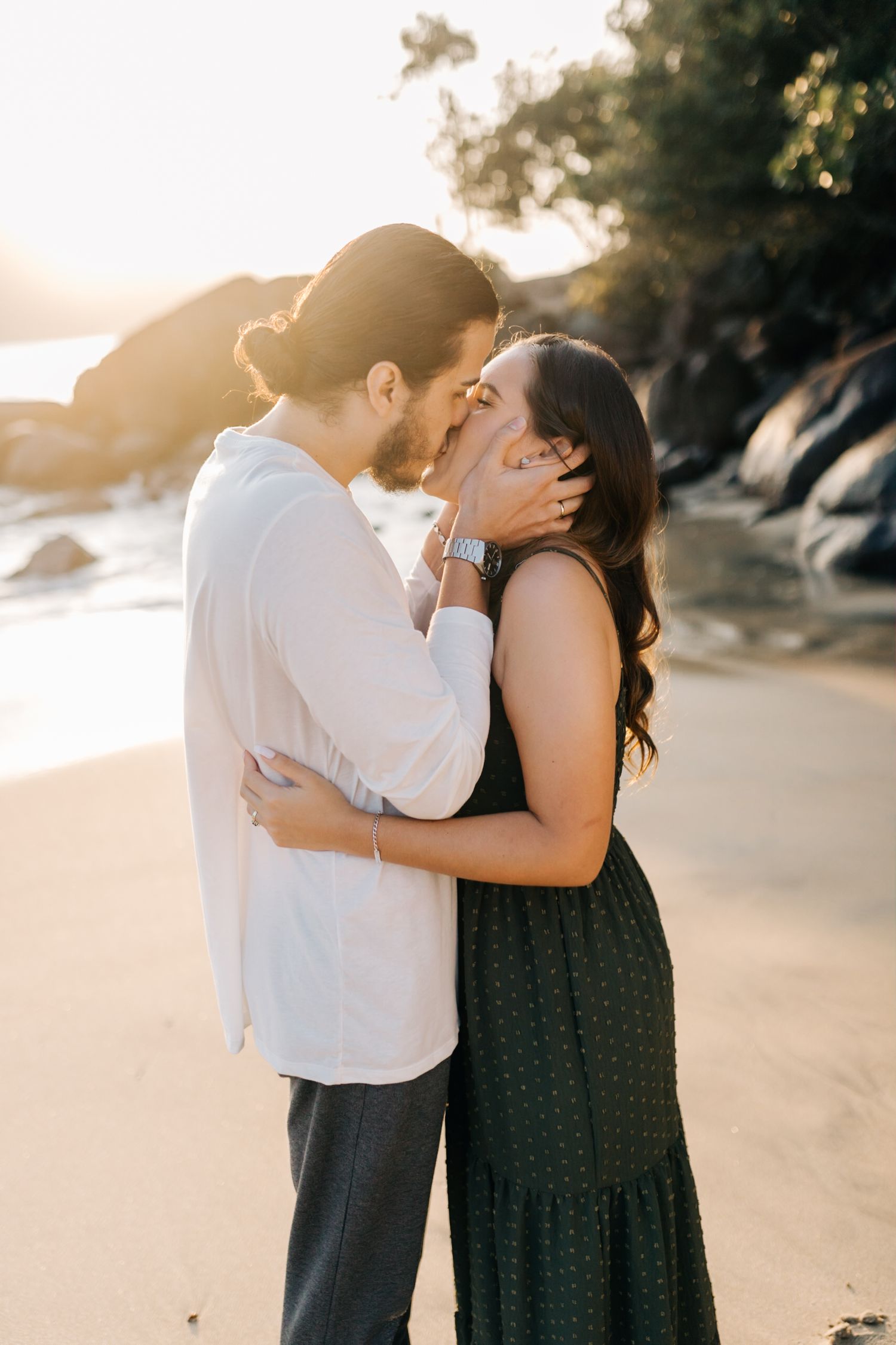 Intimate embrace captured on beach with sunlit rocks in background.