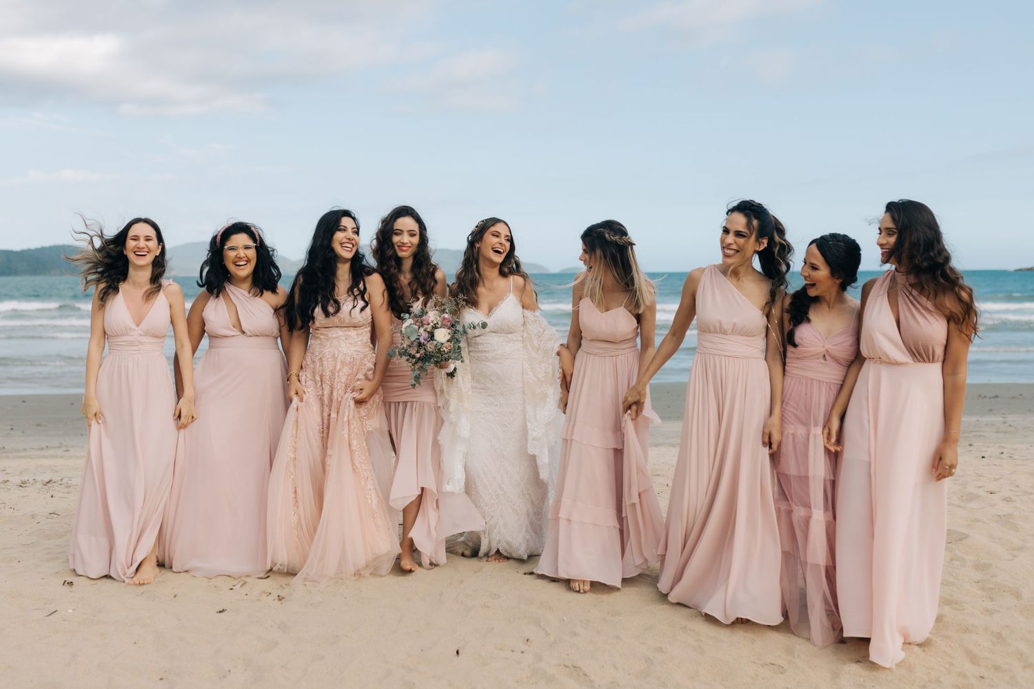 Bridesmaids in flowing pink dresses pose together on a sandy beach with ocean backdrop.