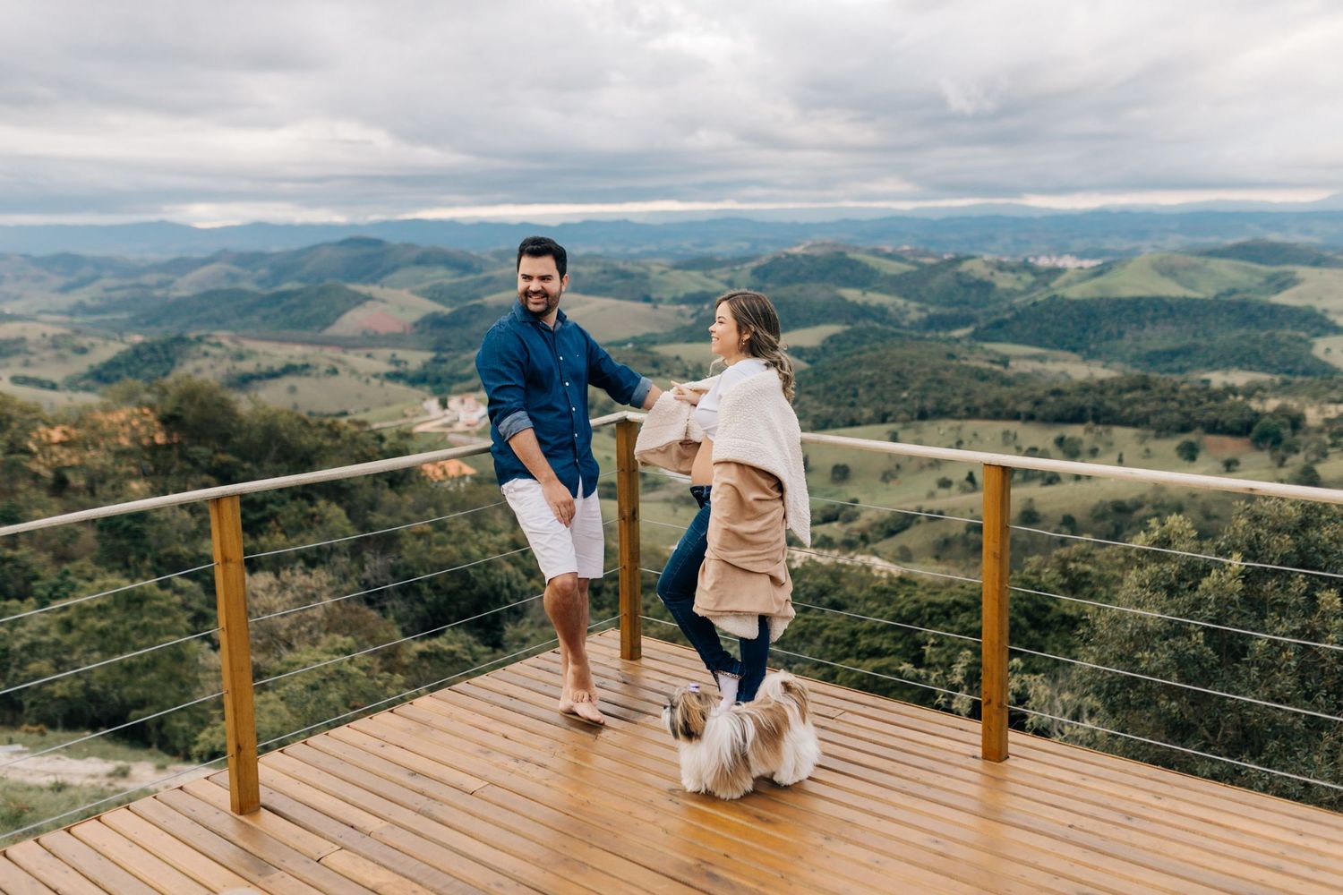 Two people and a dog stand on a mountaintop viewing deck overlooking rolling hills and valleys.