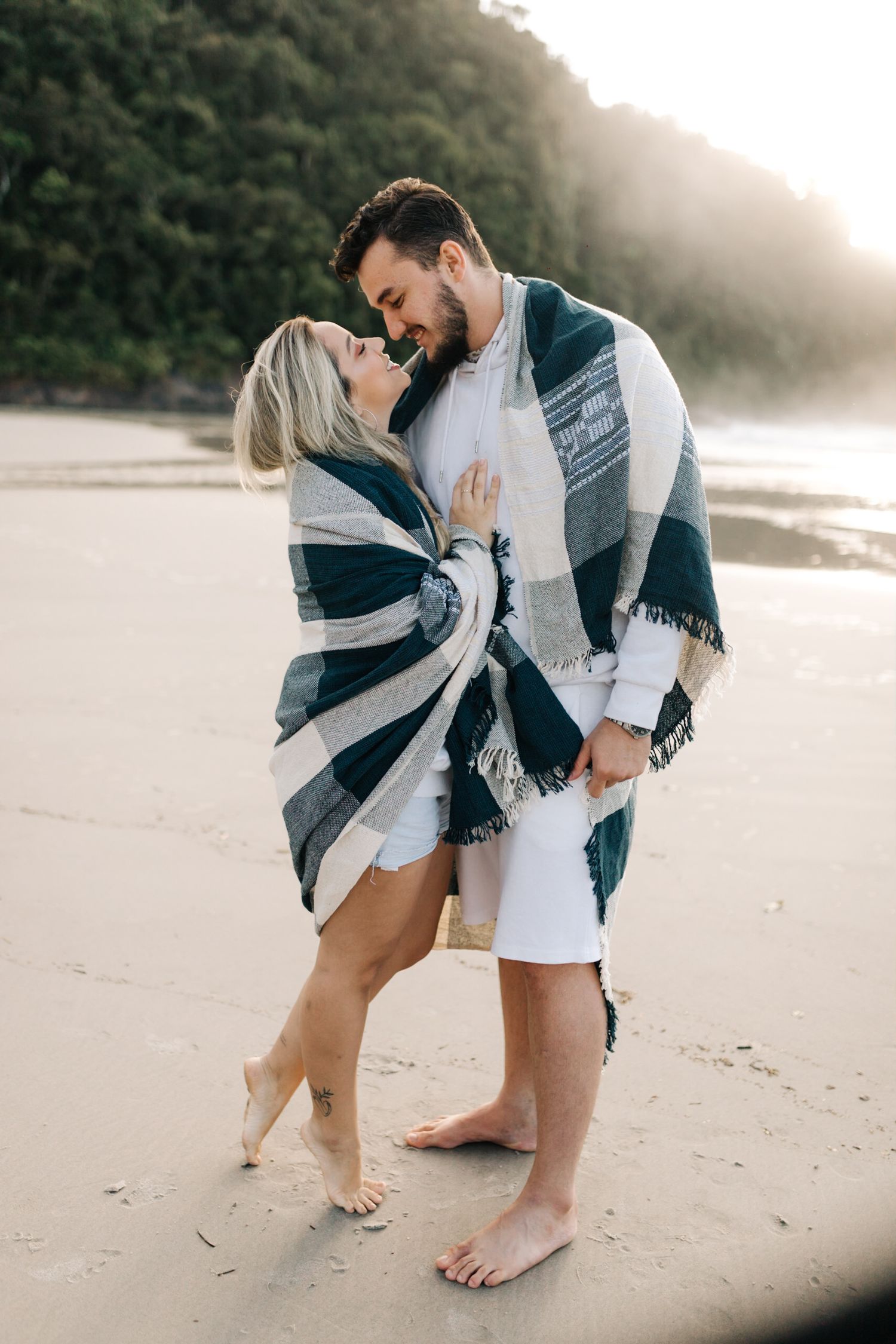 A couple wrapped in a striped blanket shares an intimate moment on a beach at sunset.
