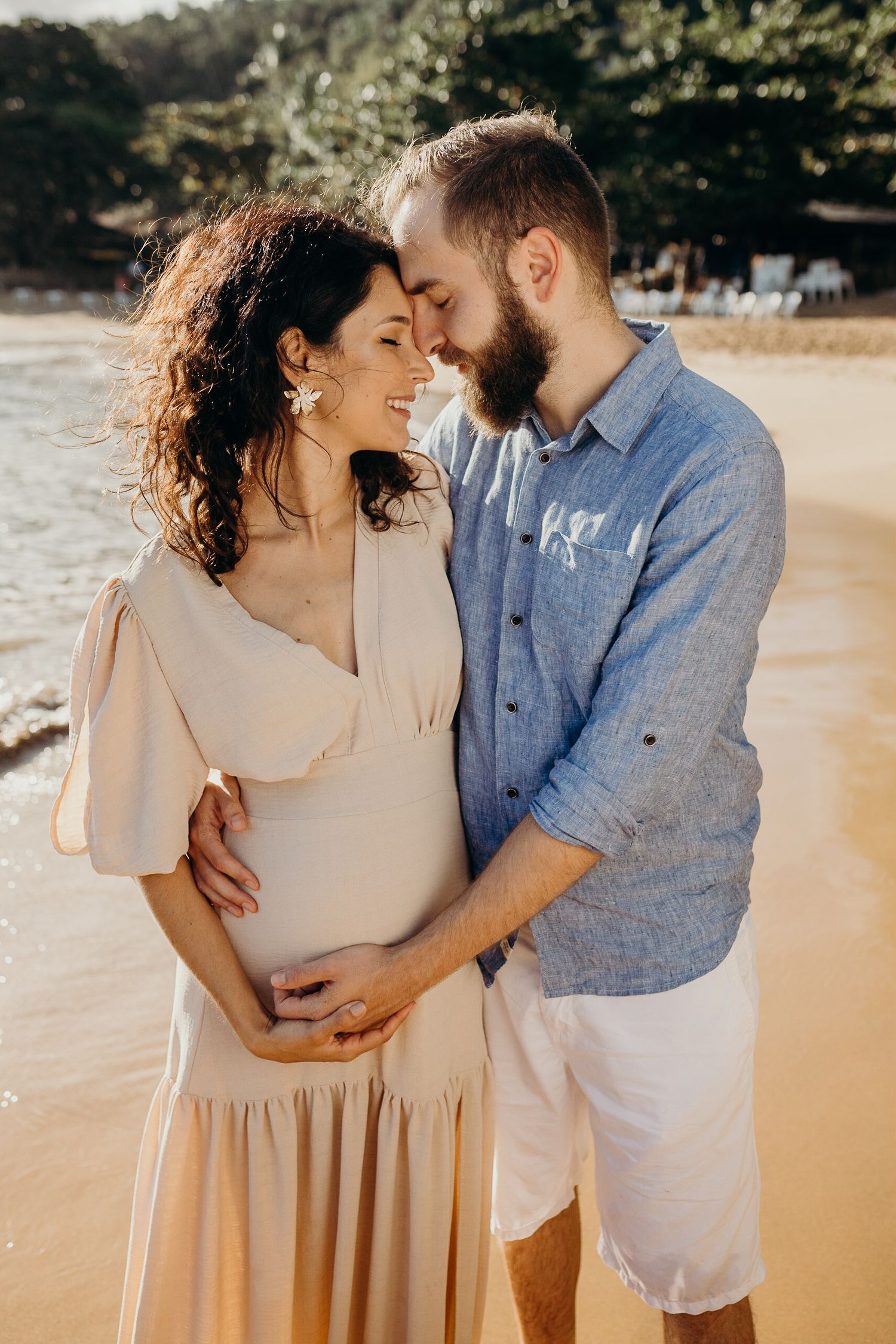 An expectant couple embraces on a beach at sunset in matching casual attire.