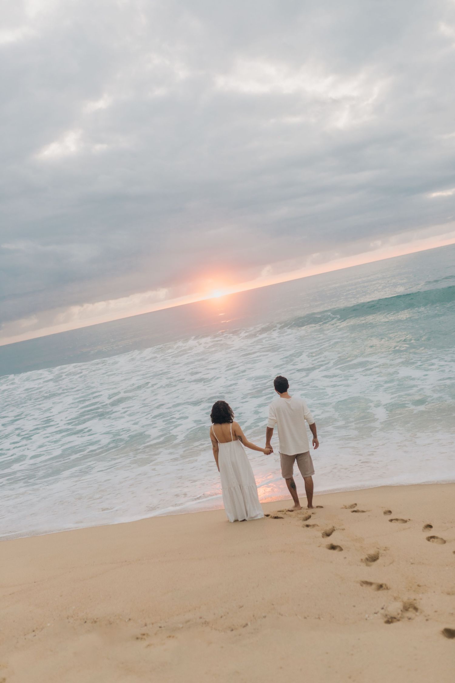 Two people in white attire stand on the beach watching the sun set over turquoise waters.