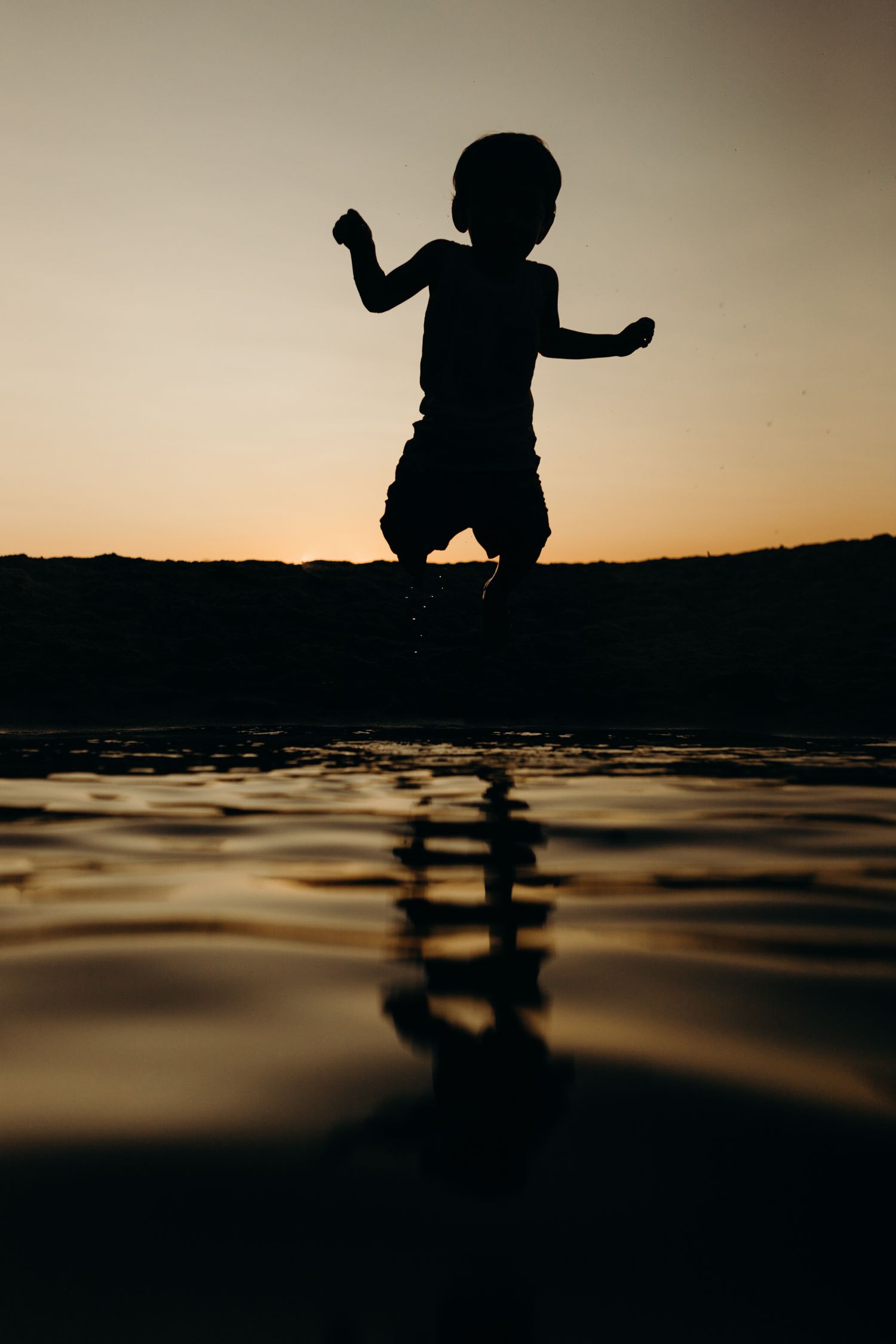 Silhouette of child jumping over water at sunset creating reflection on surface.