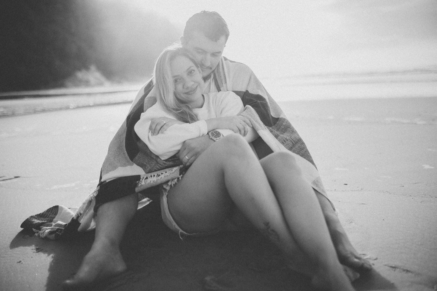 A couple wrapped in blankets enjoying an intimate moment on the shoreline.