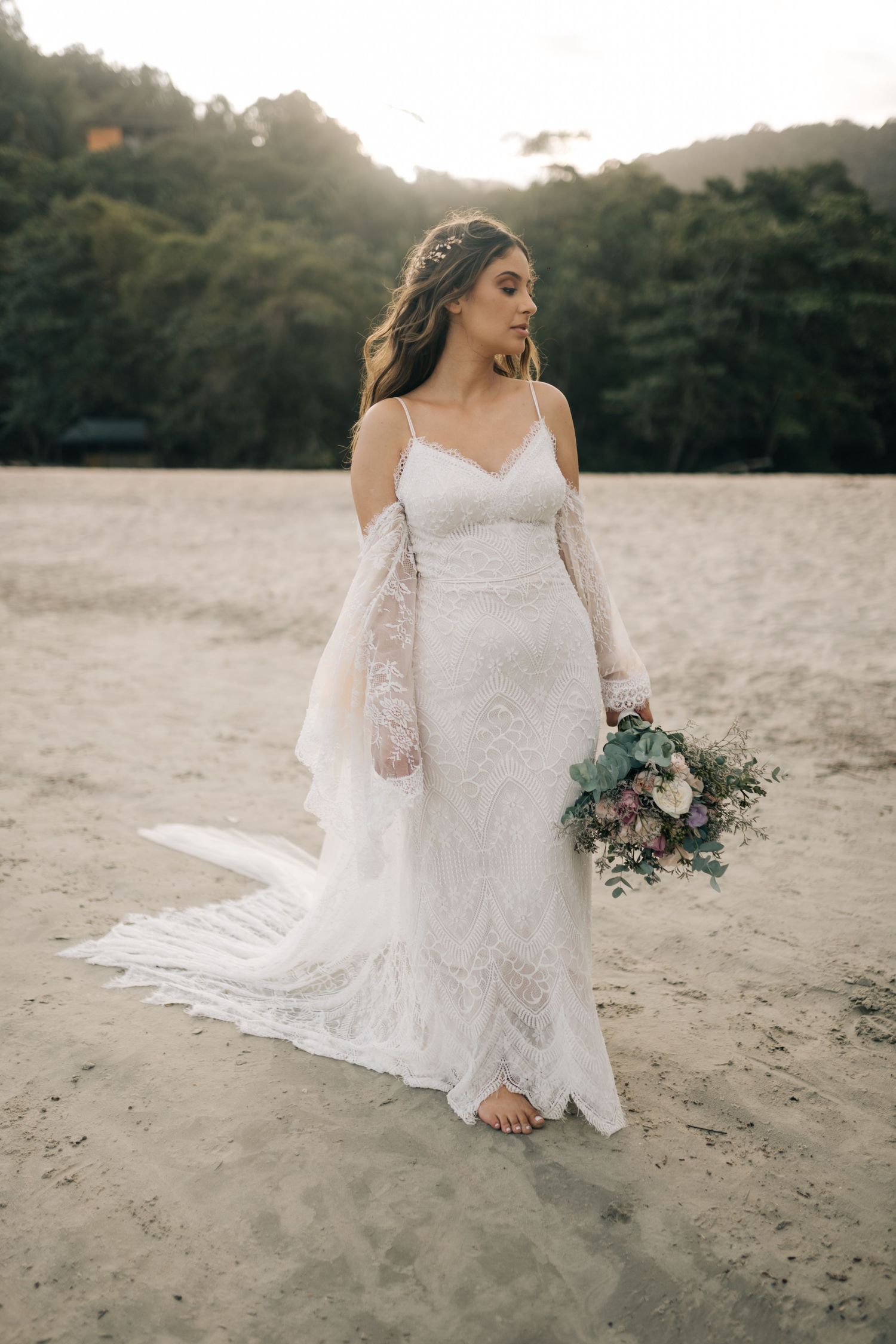 Bride in flowing lace wedding dress holding bouquet on beach at dusk.