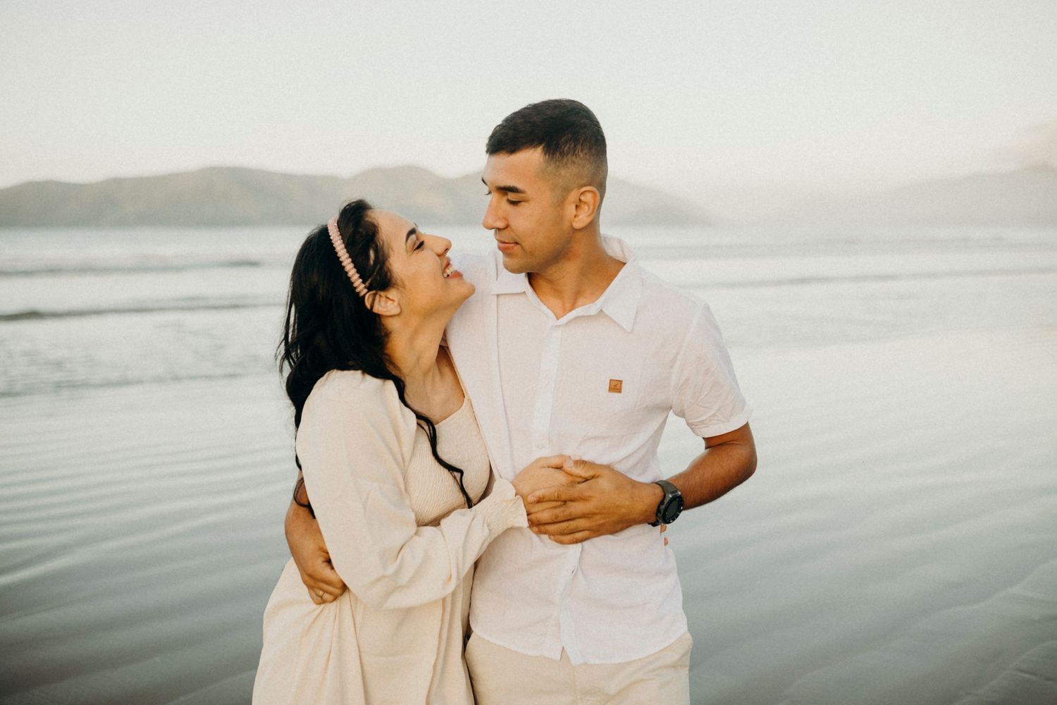 Romantic couple sharing an intimate moment on beach shore with mountains in the distance.