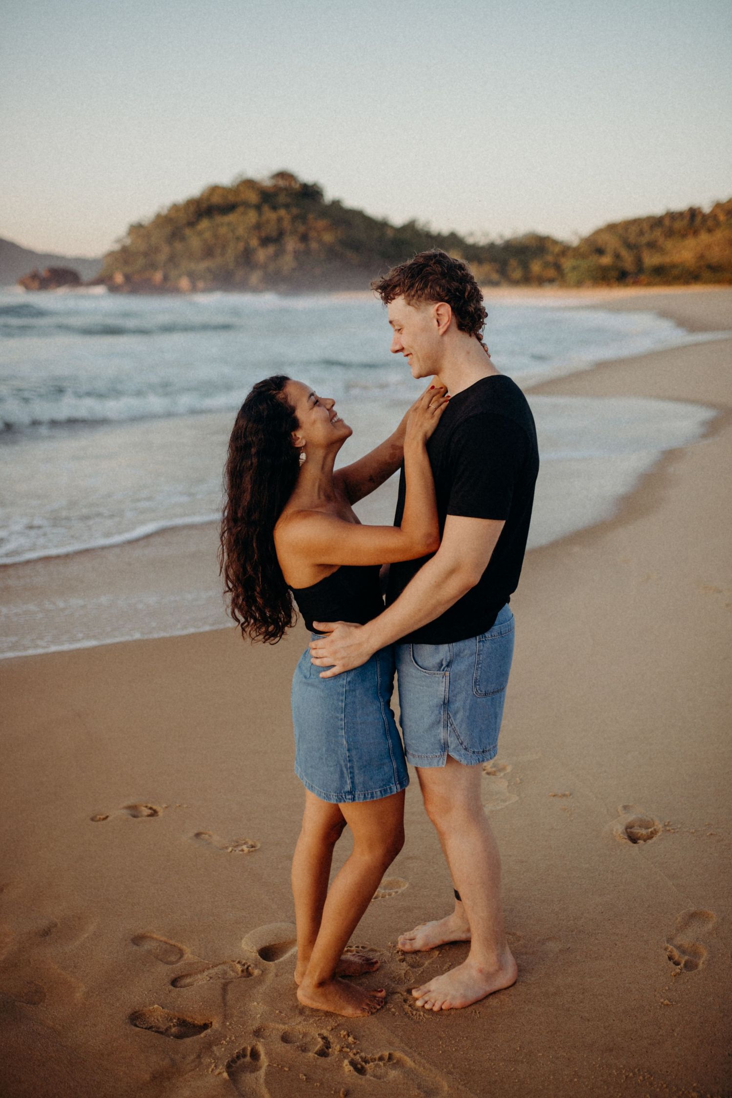 Playful couple sharing a romantic moment on beach with coastal scenery in background.