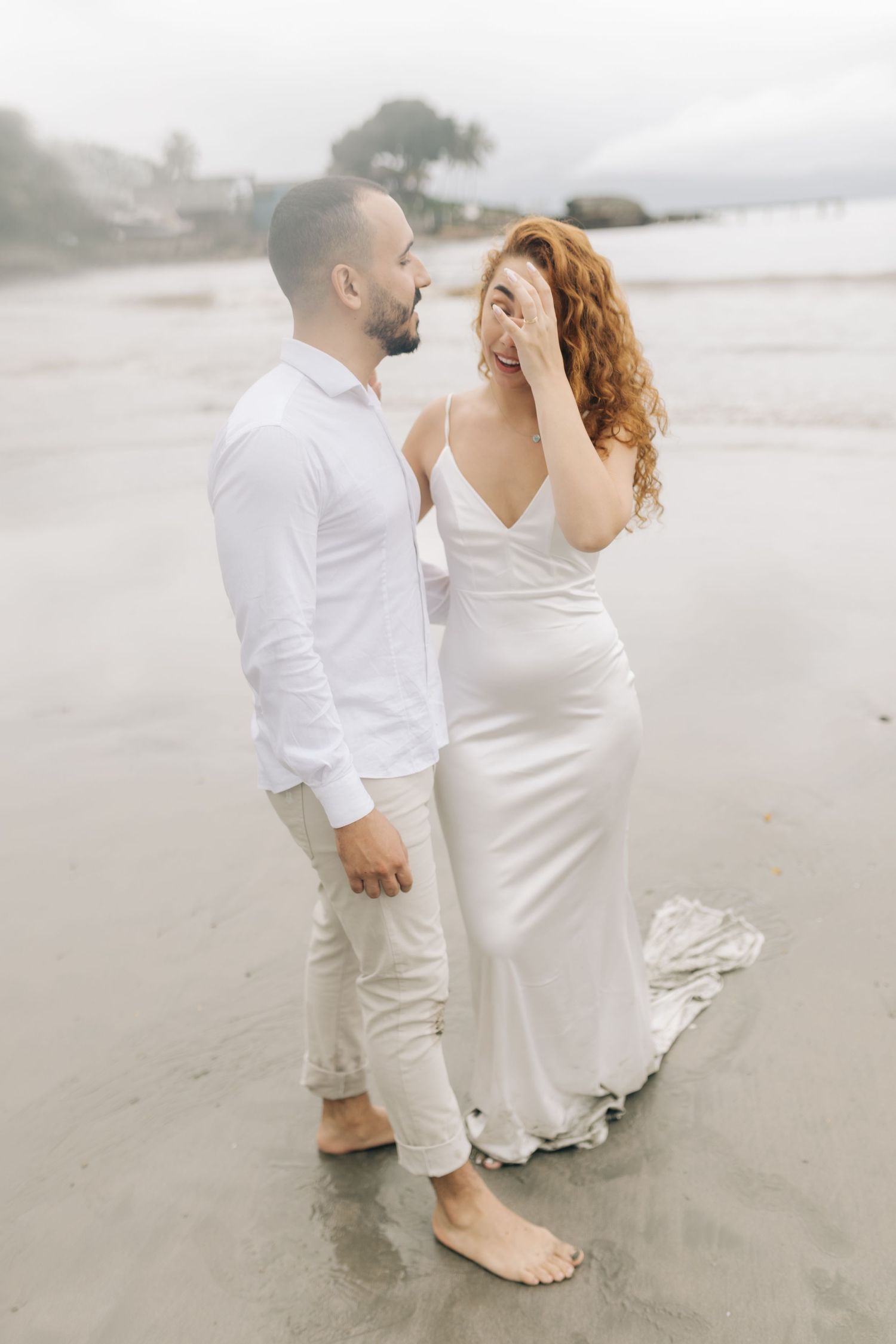 A couple laughs together during their beach photoshoot wearing coordinated white attire.