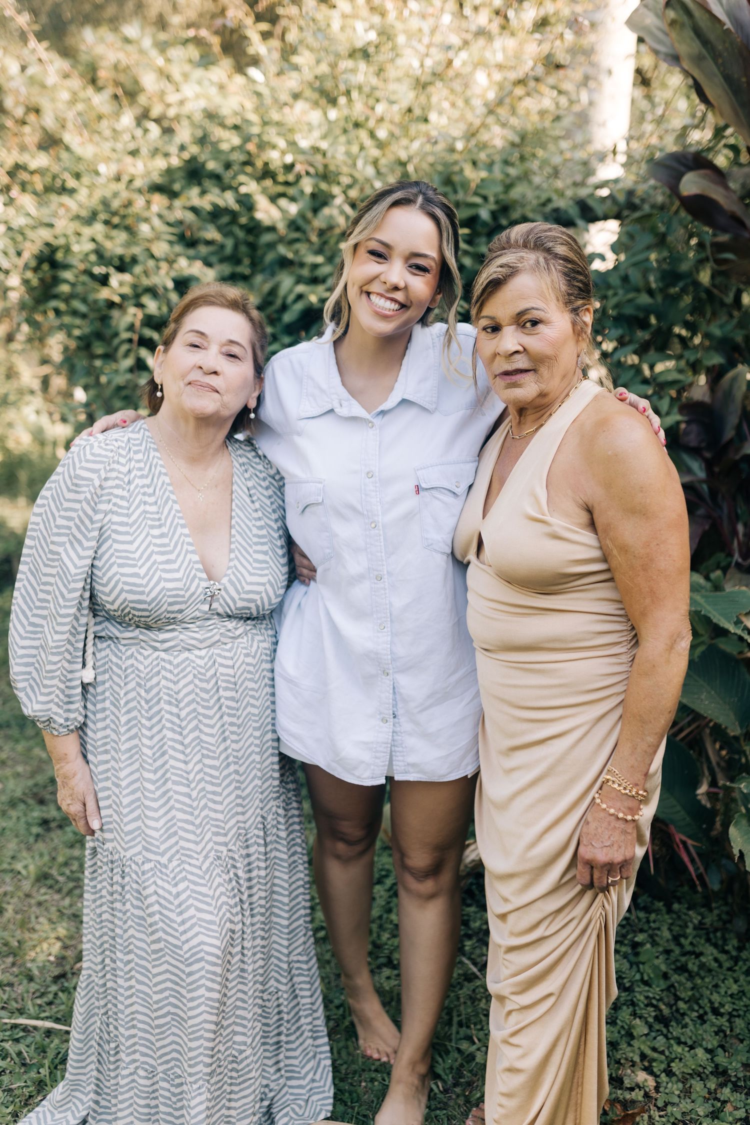 Three friends pose together outdoors in casual summer outfits against a green foliage backdrop.