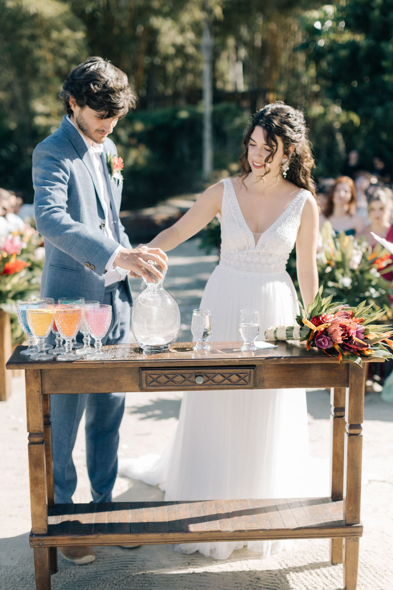 Wedding couple stands at a wooden table with glassware and drink stations at their outdoor reception.