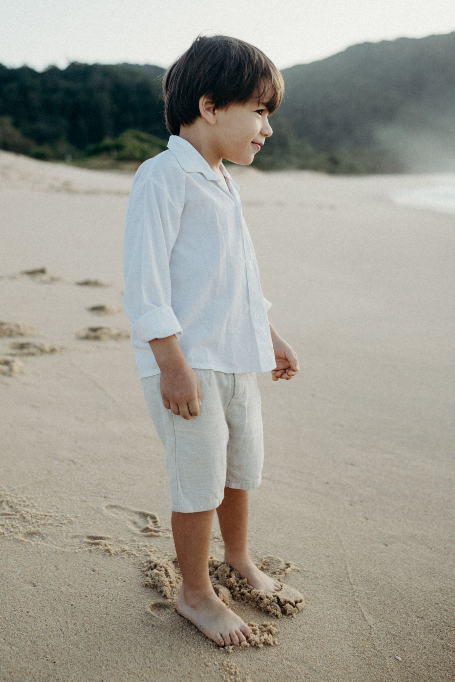 A young child in white attire standing barefoot on wet sand at the beach.