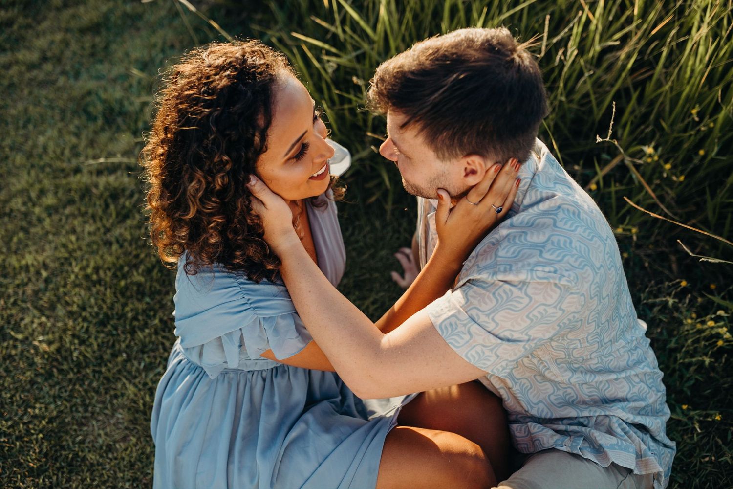 Couple sharing tender moment while sitting in tall grass during sunset.