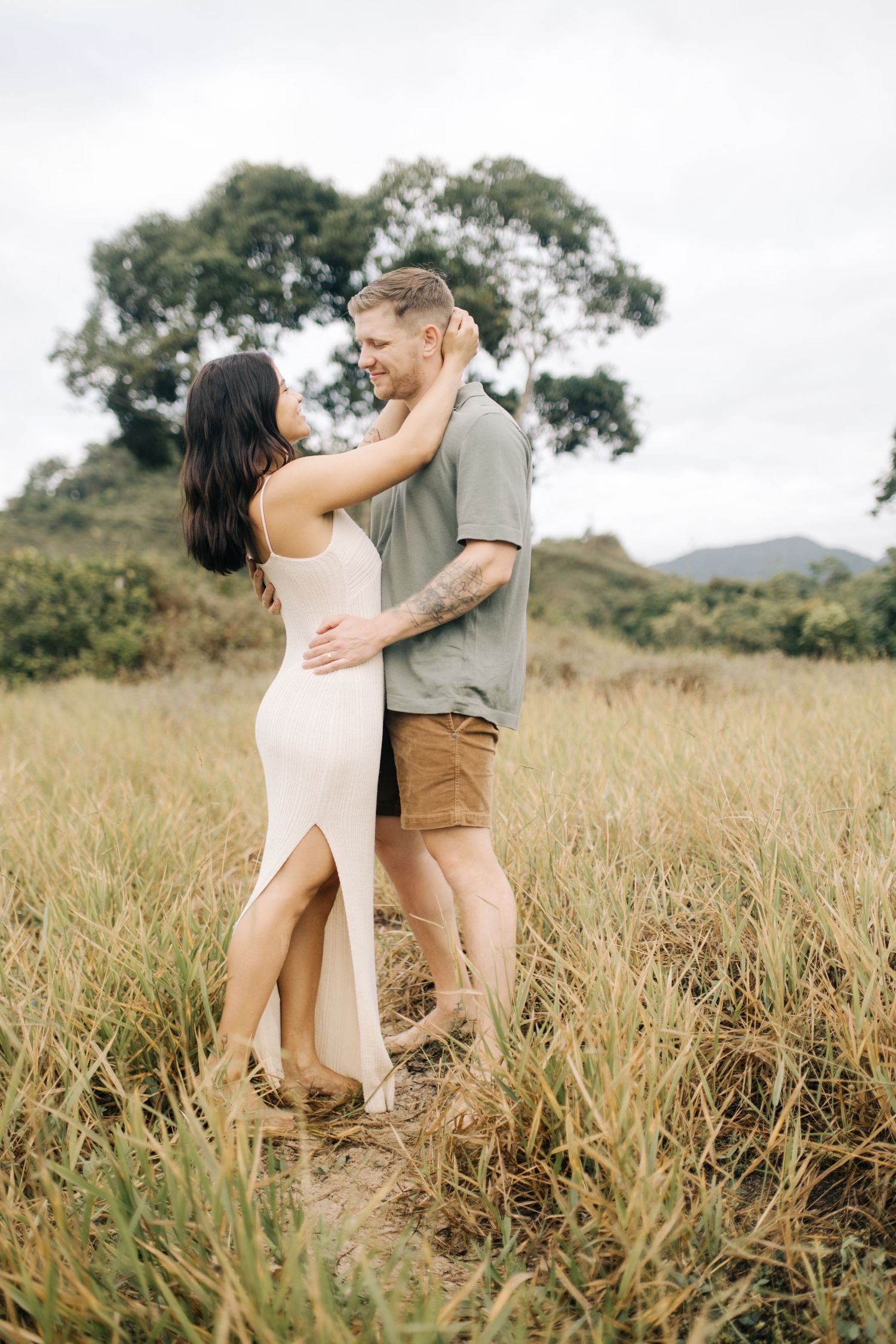 A romantic couple shares intimate moments together in a grassy field with mountains in the background.