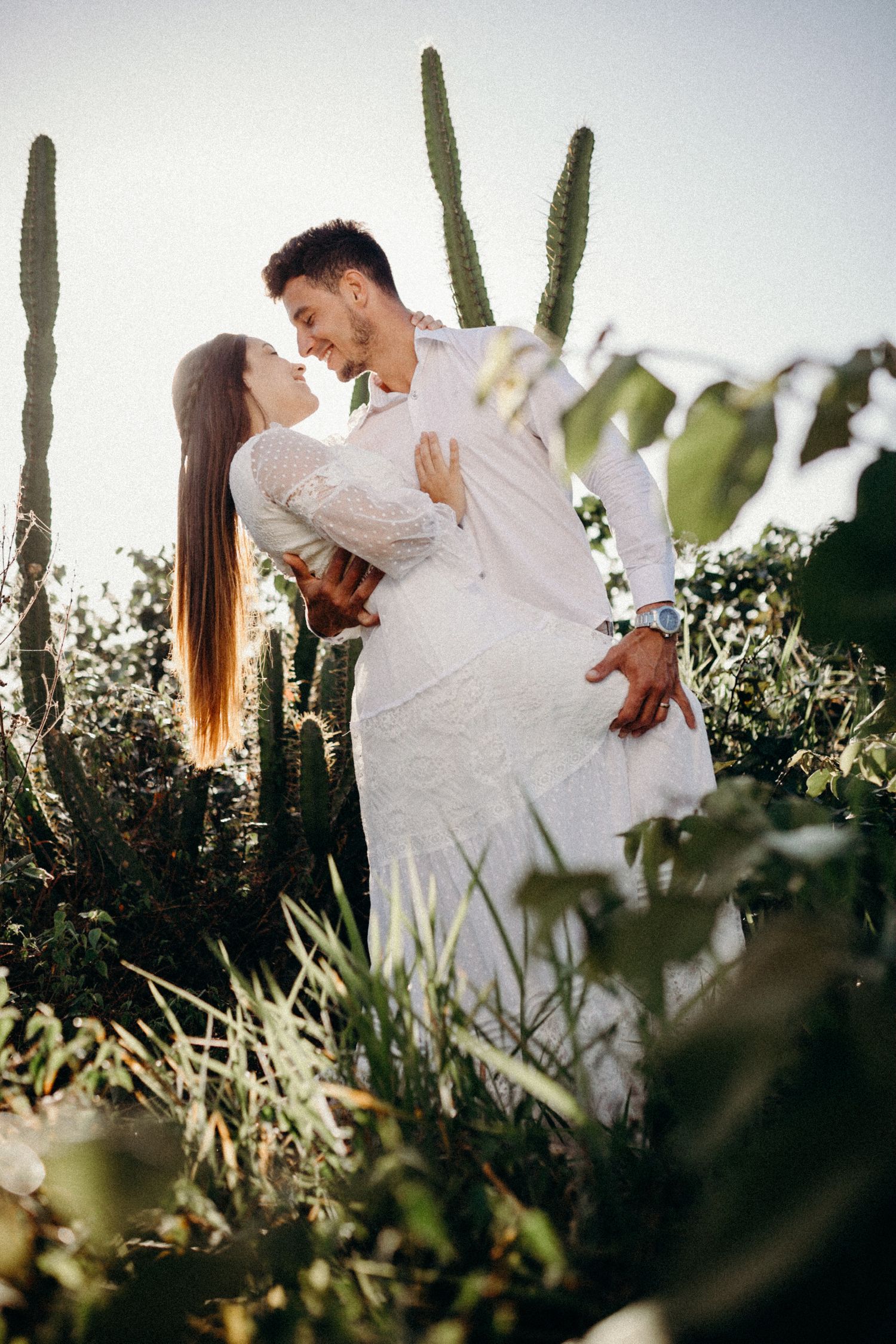 Couple in light clothing share intimate moment in desert landscape with towering saguaro cacti.