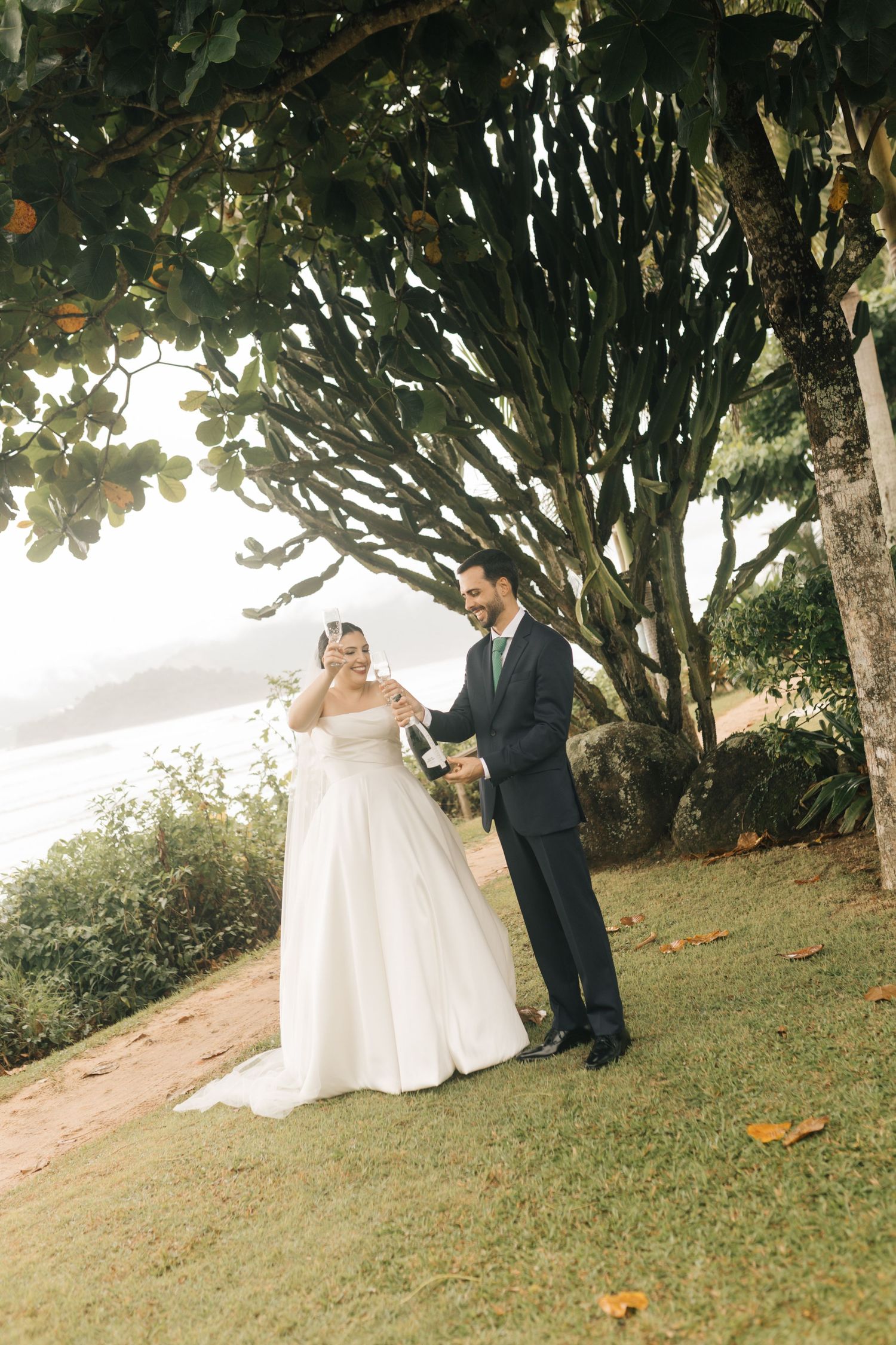 Newlyweds share a moment under a large tree on a scenic oceanside cliff.