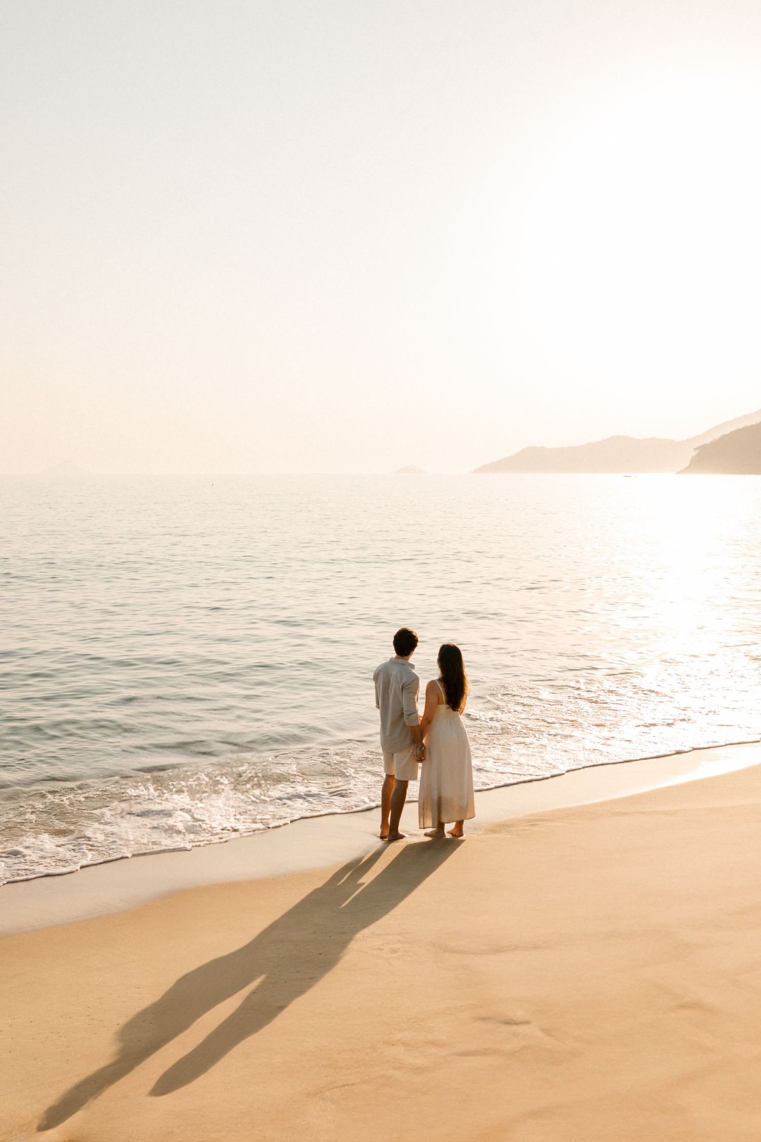 Romantic silhouette of a couple standing on the beach during golden hour with long shadows cast on sand.