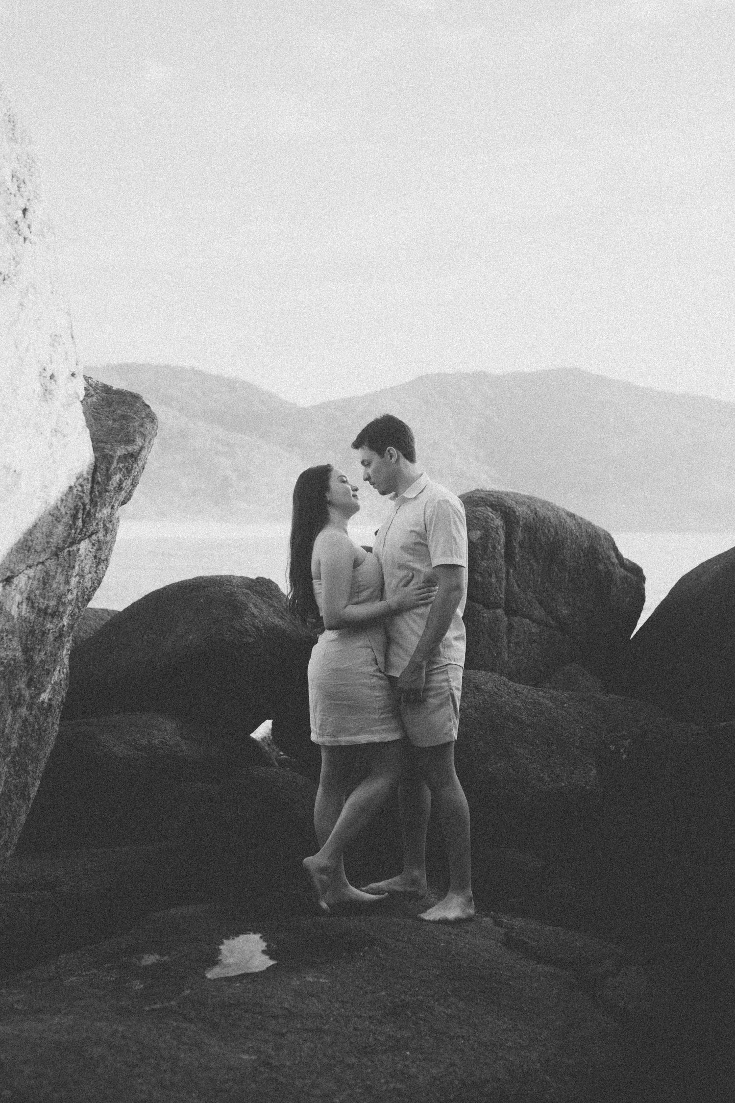 Black and white photo of couple embracing among coastal rocks with mountains in background.