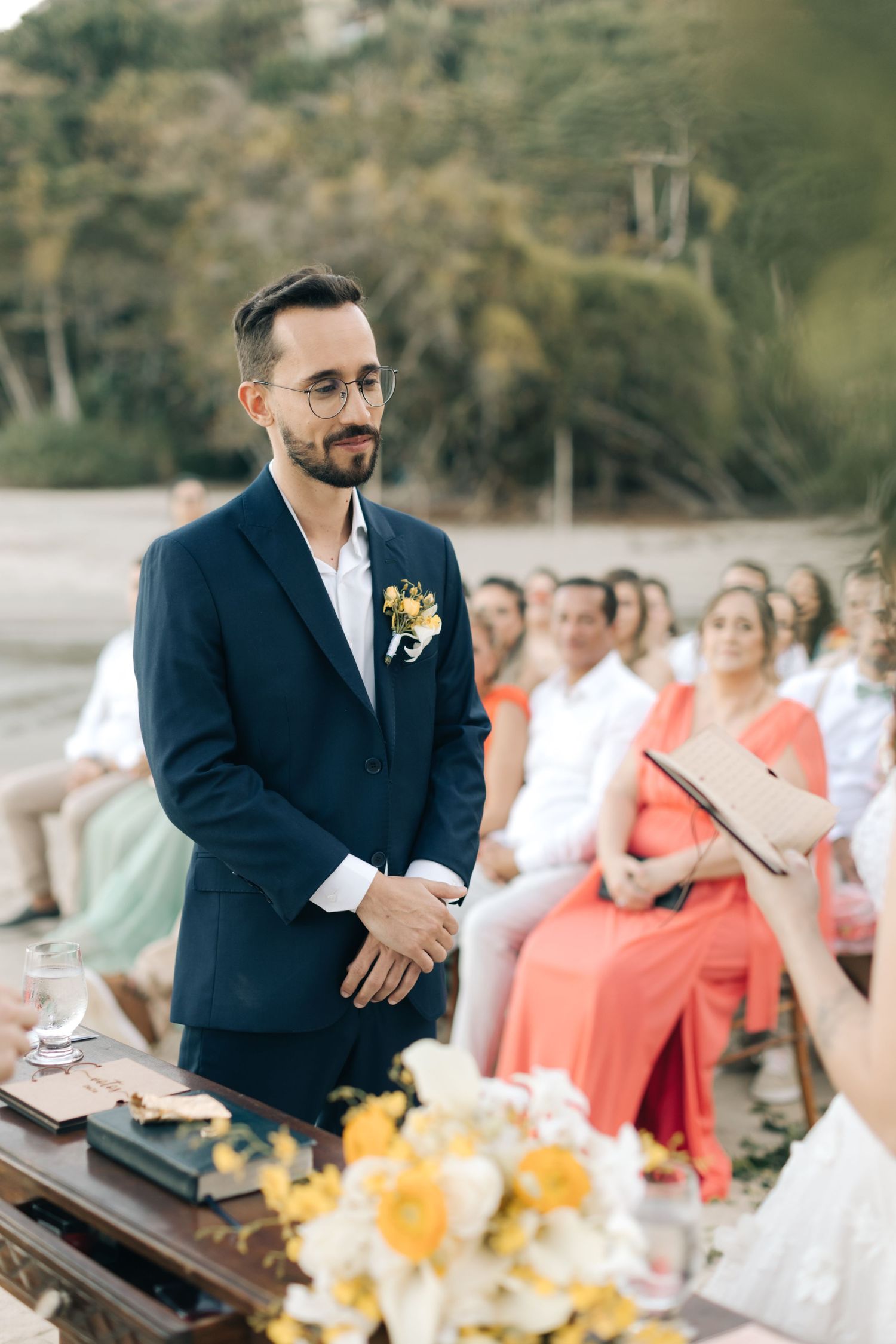 Smiling officiant in navy suit stands before wedding party during outdoor ceremony.