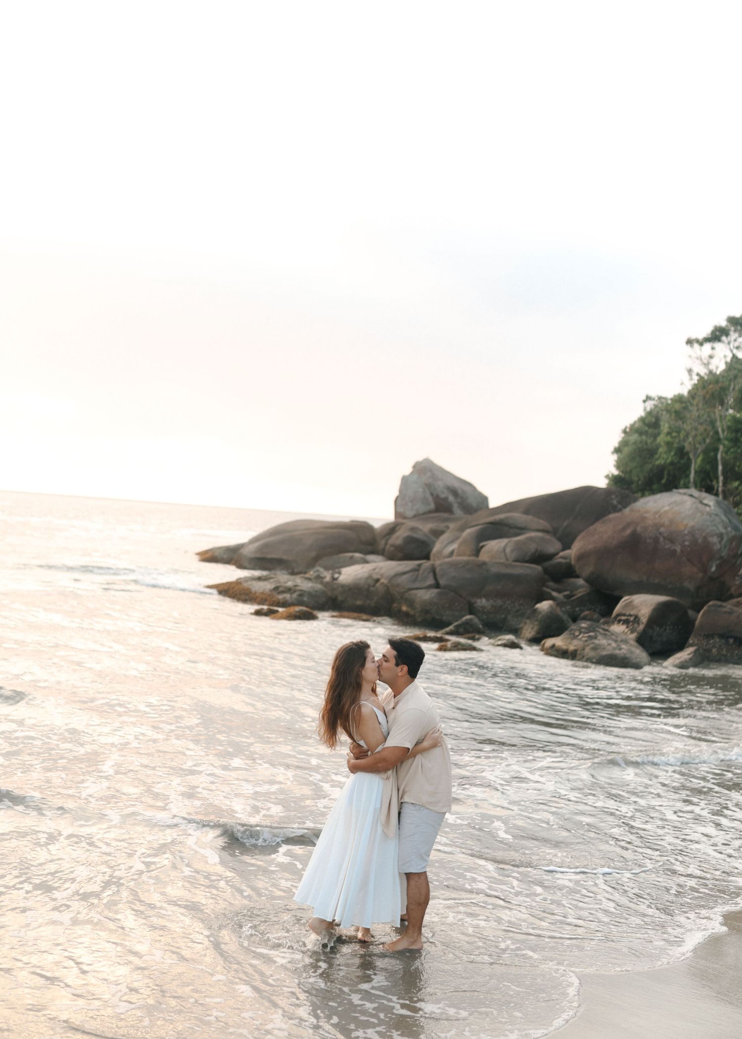 Couple in white outfits plays in ocean waves during beach photoshoot.