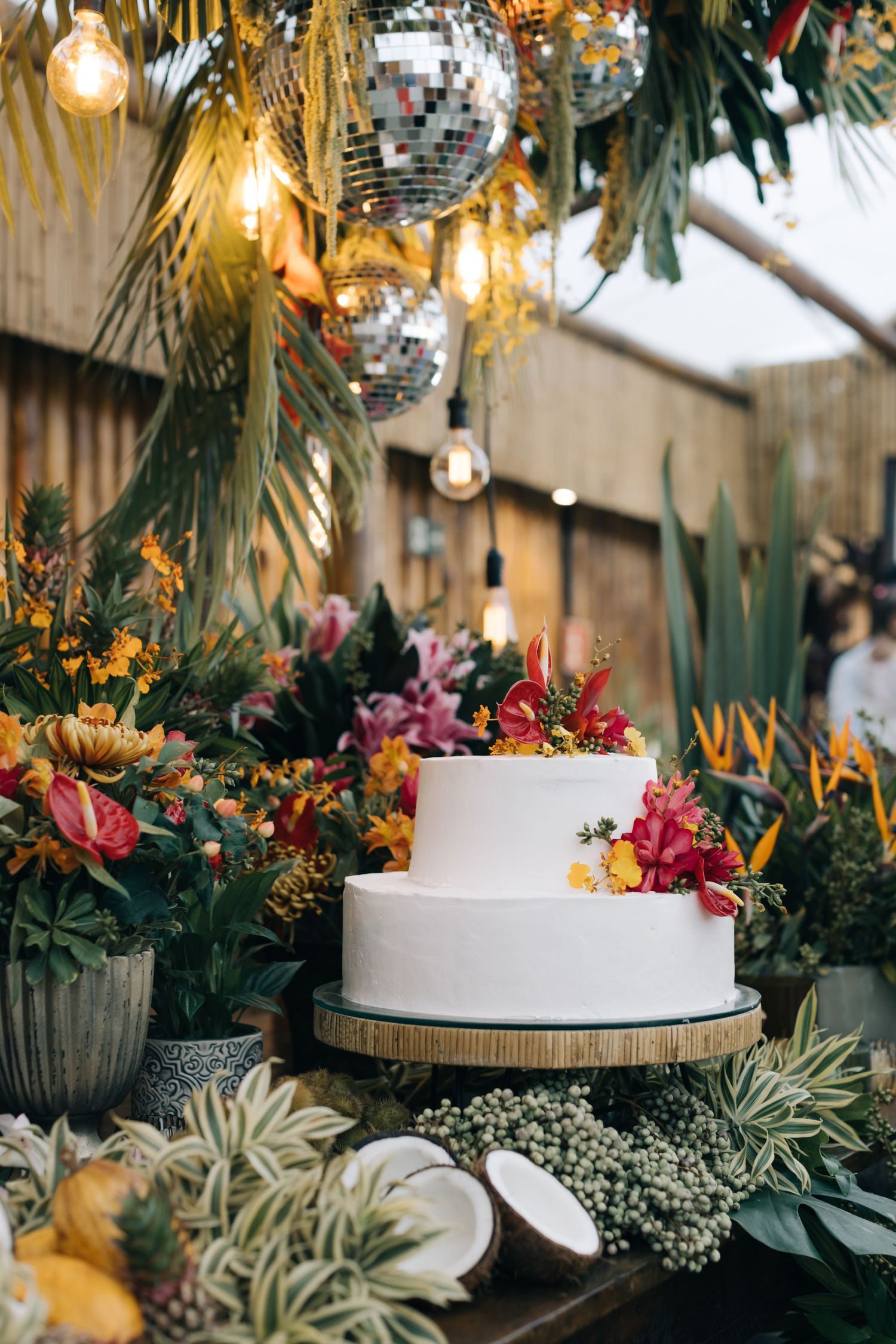 White two-tier wedding cake decorated with tropical flowers under disco balls and string lights.