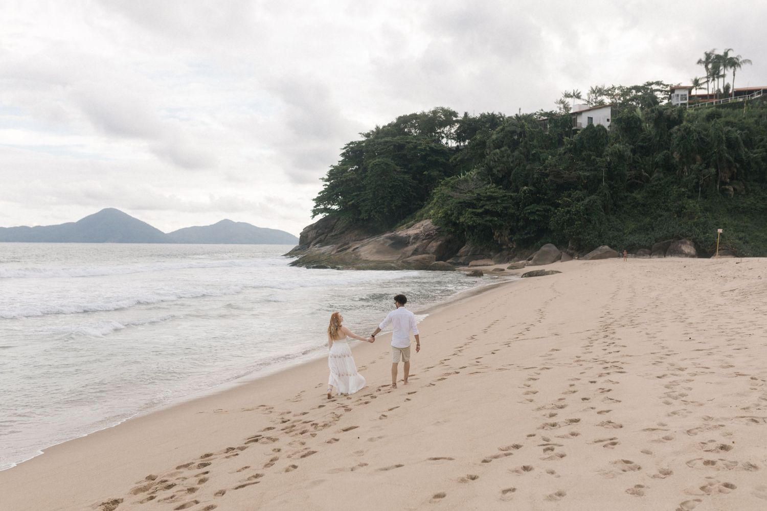 A couple walks hand in hand along a scenic beach with mountains in the background.