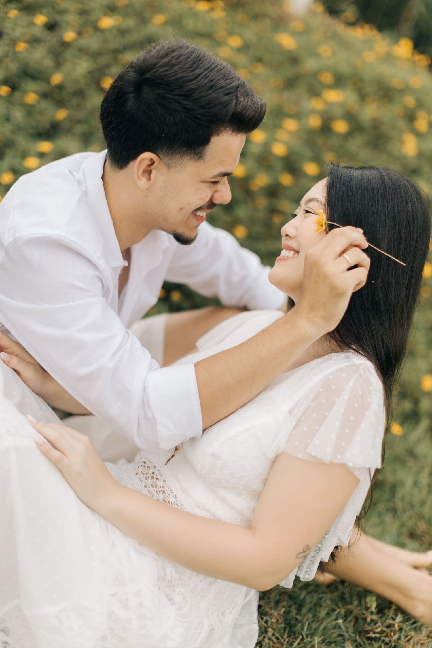Playful couple in white clothing laughing together in field of yellow flowers.