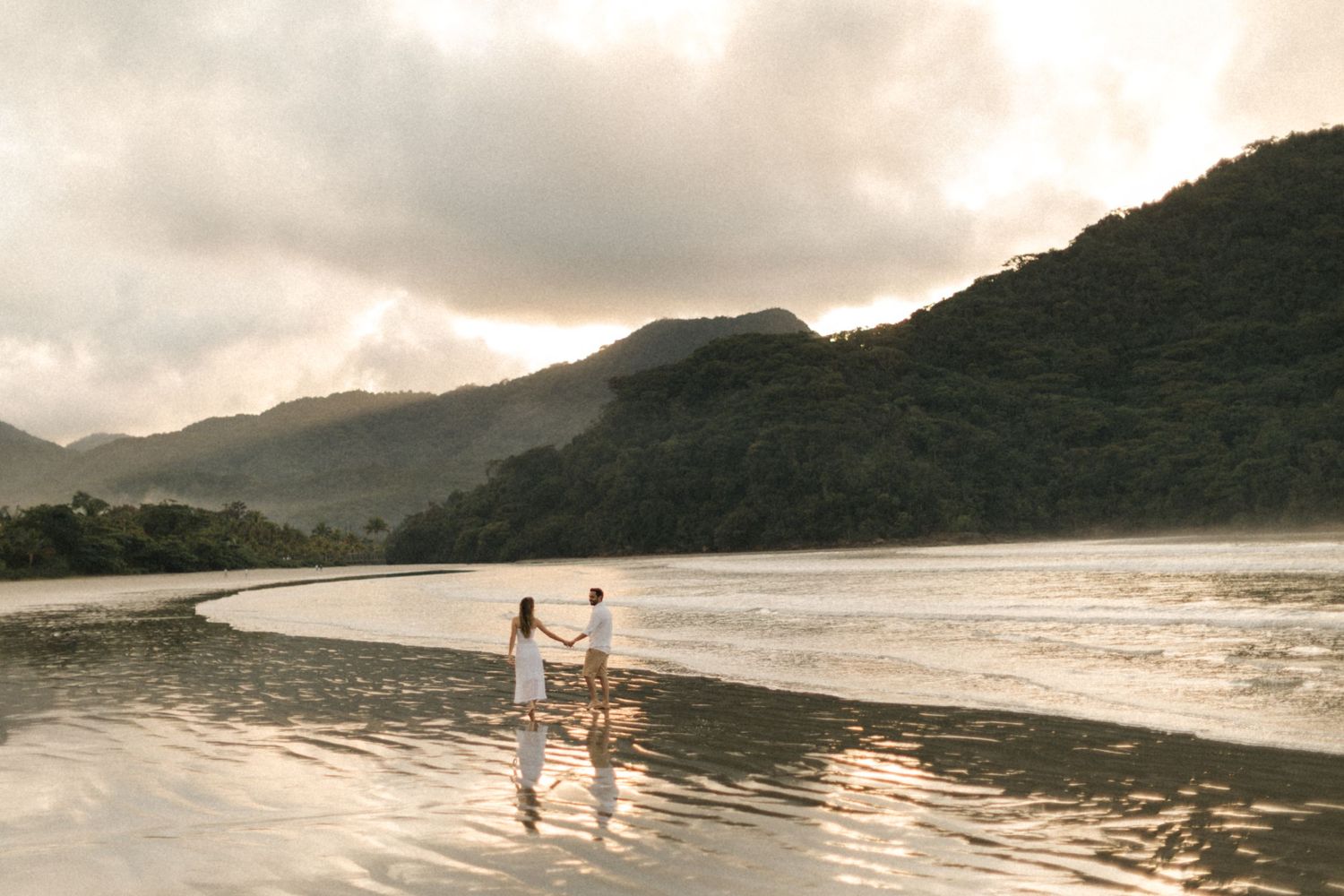Panoramic view of a couple walking along a serene beach with mountains and cloudy skies.