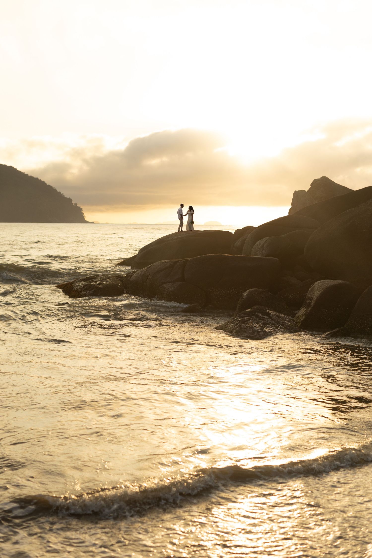 Golden sunlight illuminates silhouetted figures on rocky coastline with dramatic ocean waves crashing below.