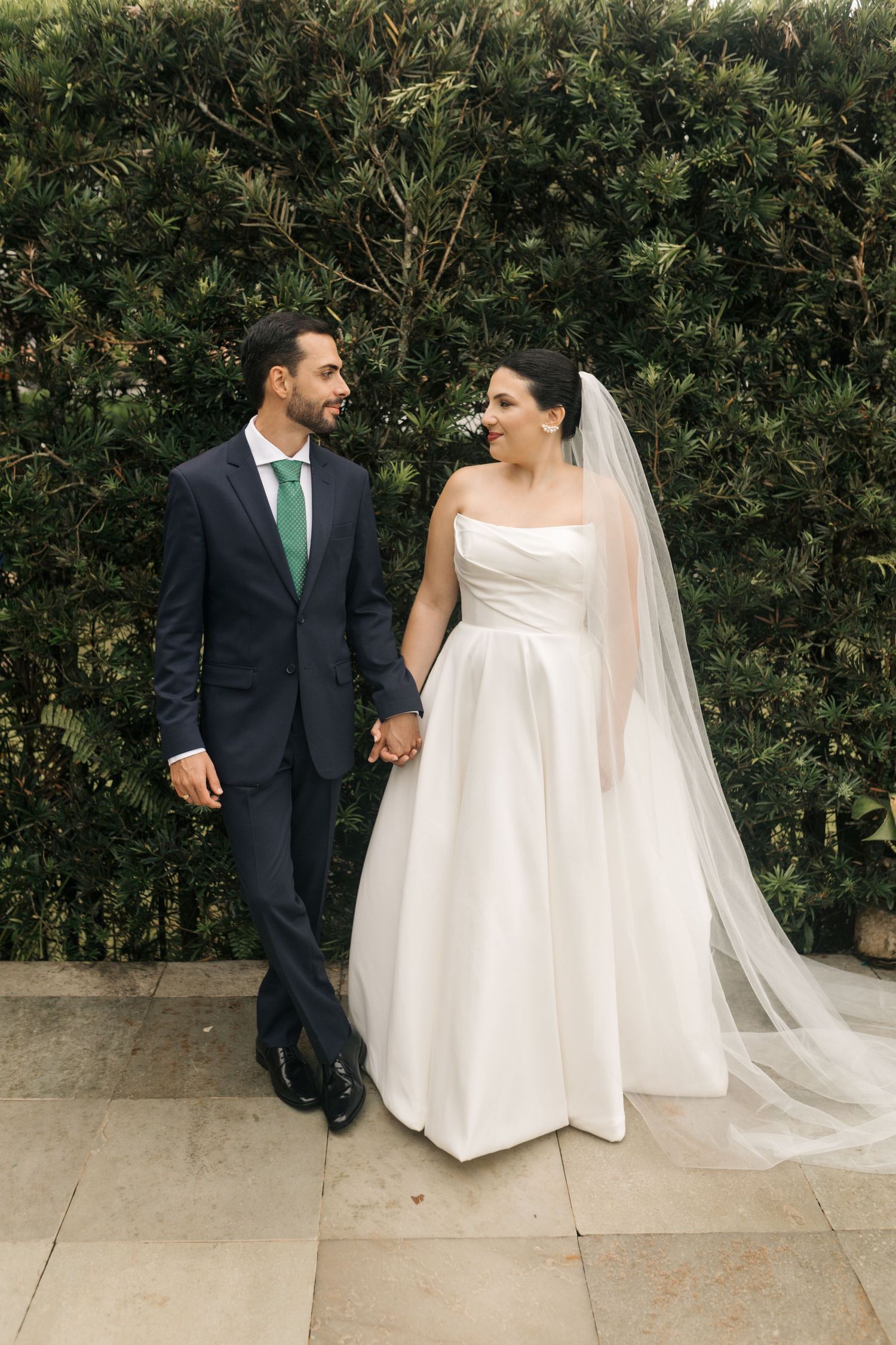 Newlyweds pose together against a lush green hedge backdrop.