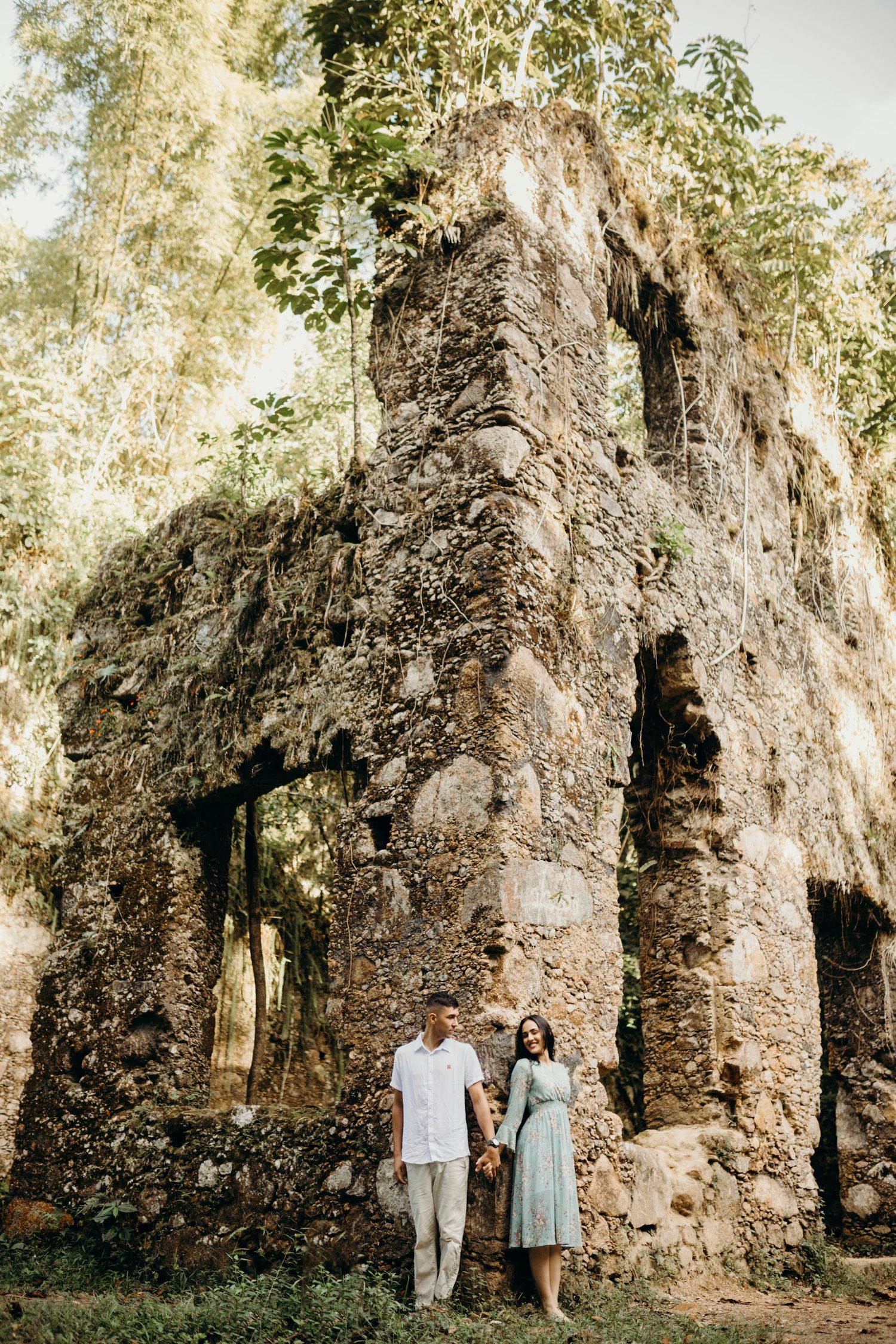 A couple poses in front of towering stone ruins surrounded by lush greenery.