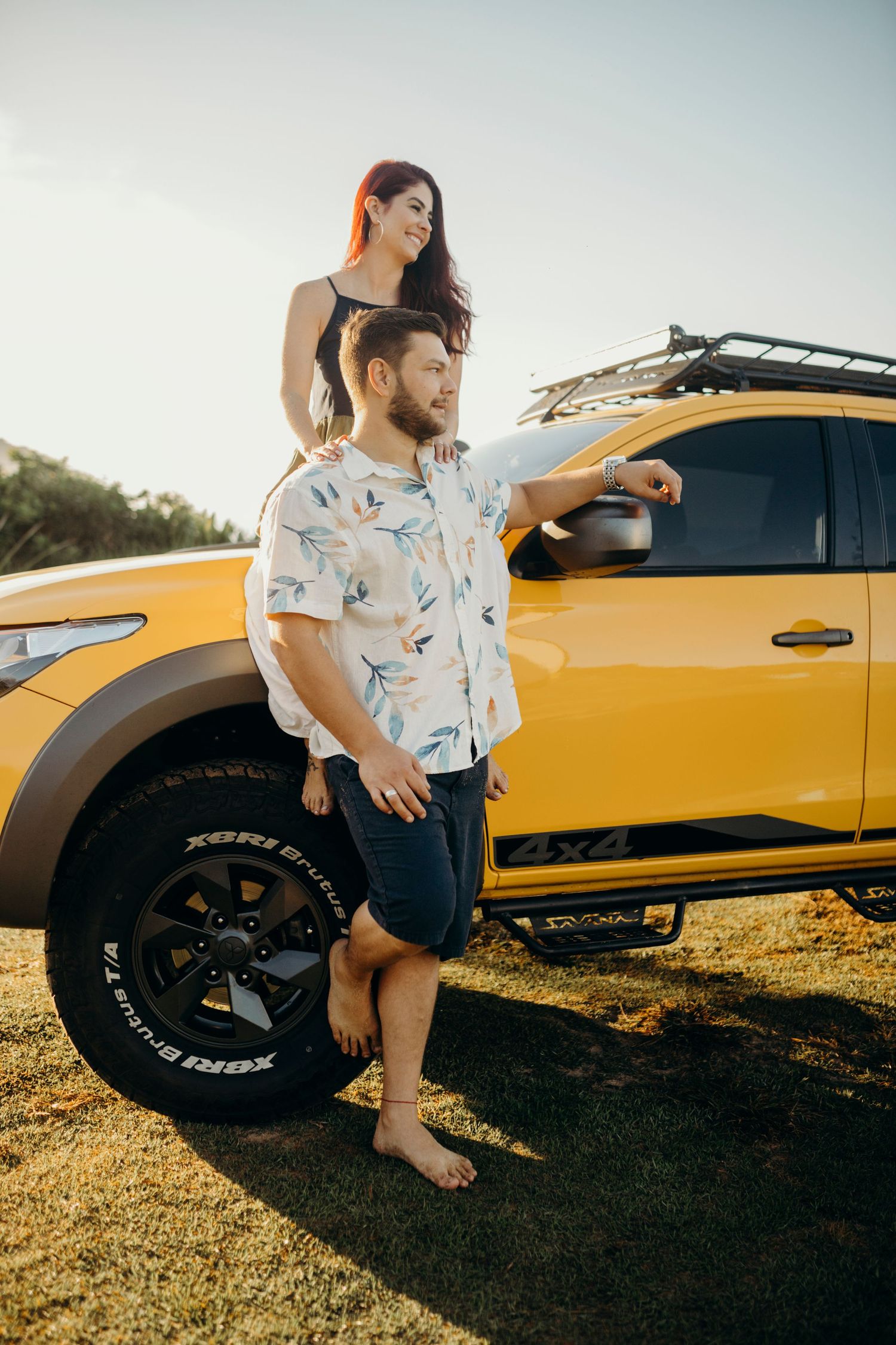Couple in casual attire posing next to yellow pickup truck at scenic coastal location.