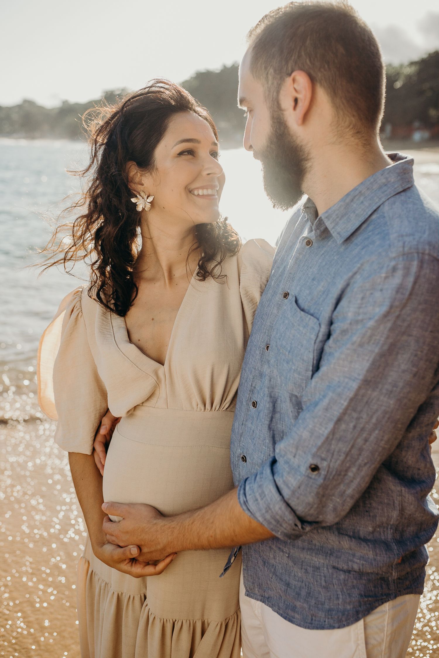 A tender beachside moment between an expecting couple in casual summer attire.