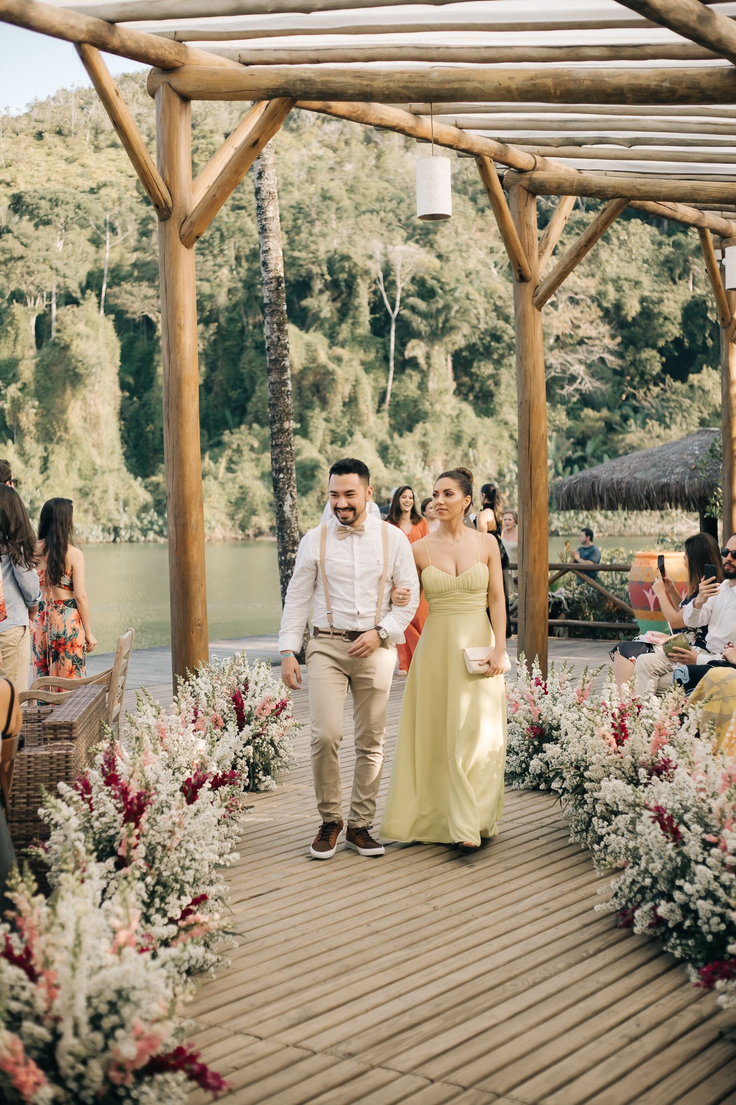 A couple walks down a wooden aisle decorated with pink and white flowers under a rustic pergola by a lake.