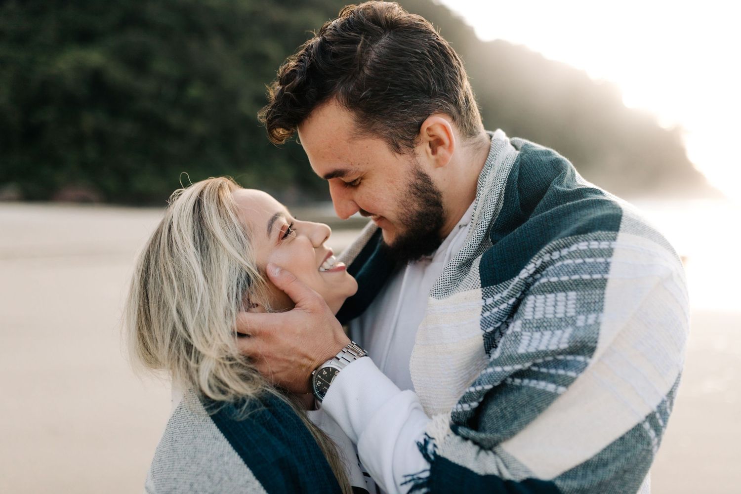 A joyful couple shares a tender moment while wrapped in a striped blanket at the beach.