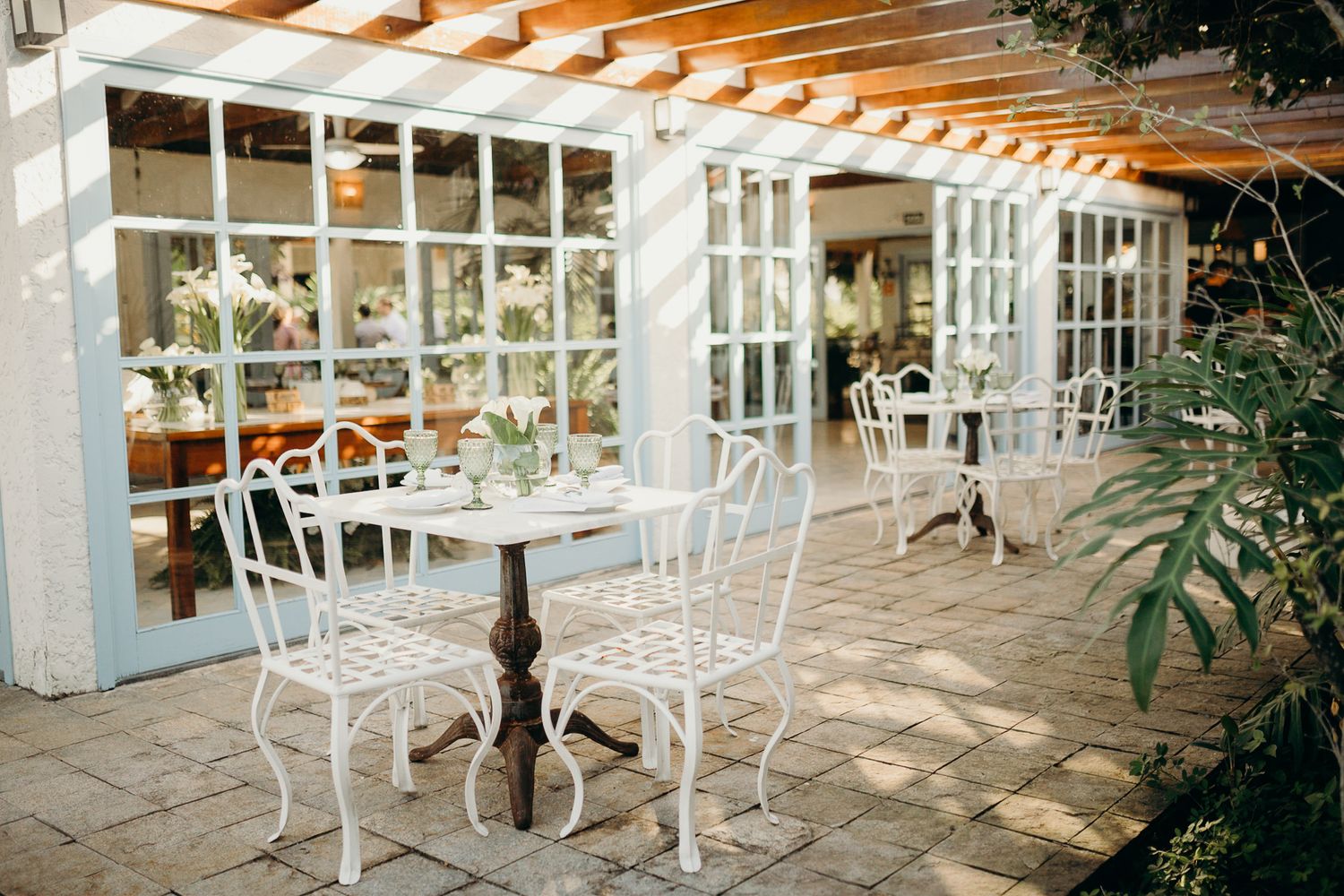 White outdoor patio furniture arranged under a wooden pergola in a garden setting.