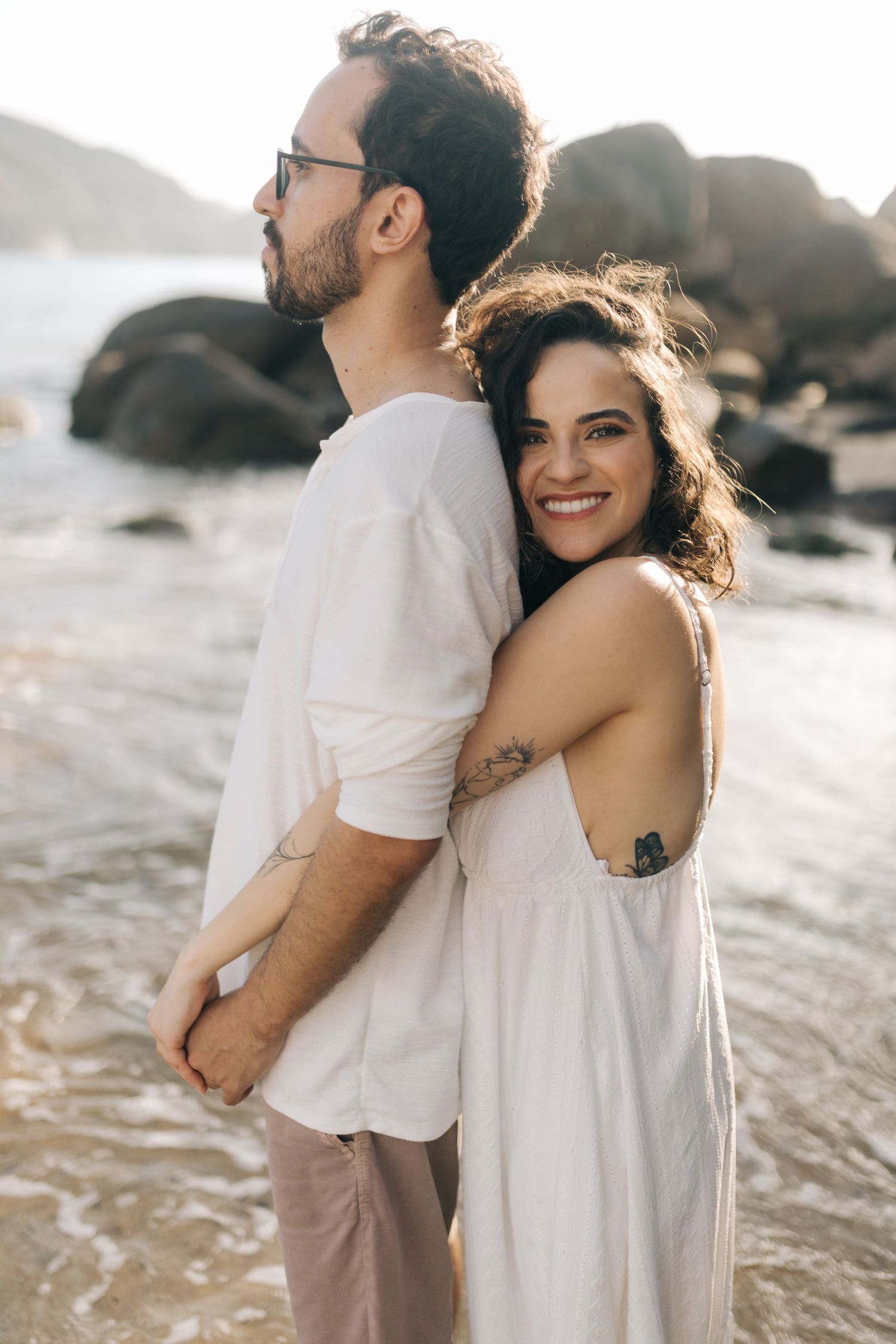 Couple in white clothing embracing on a rocky beach at sunset.