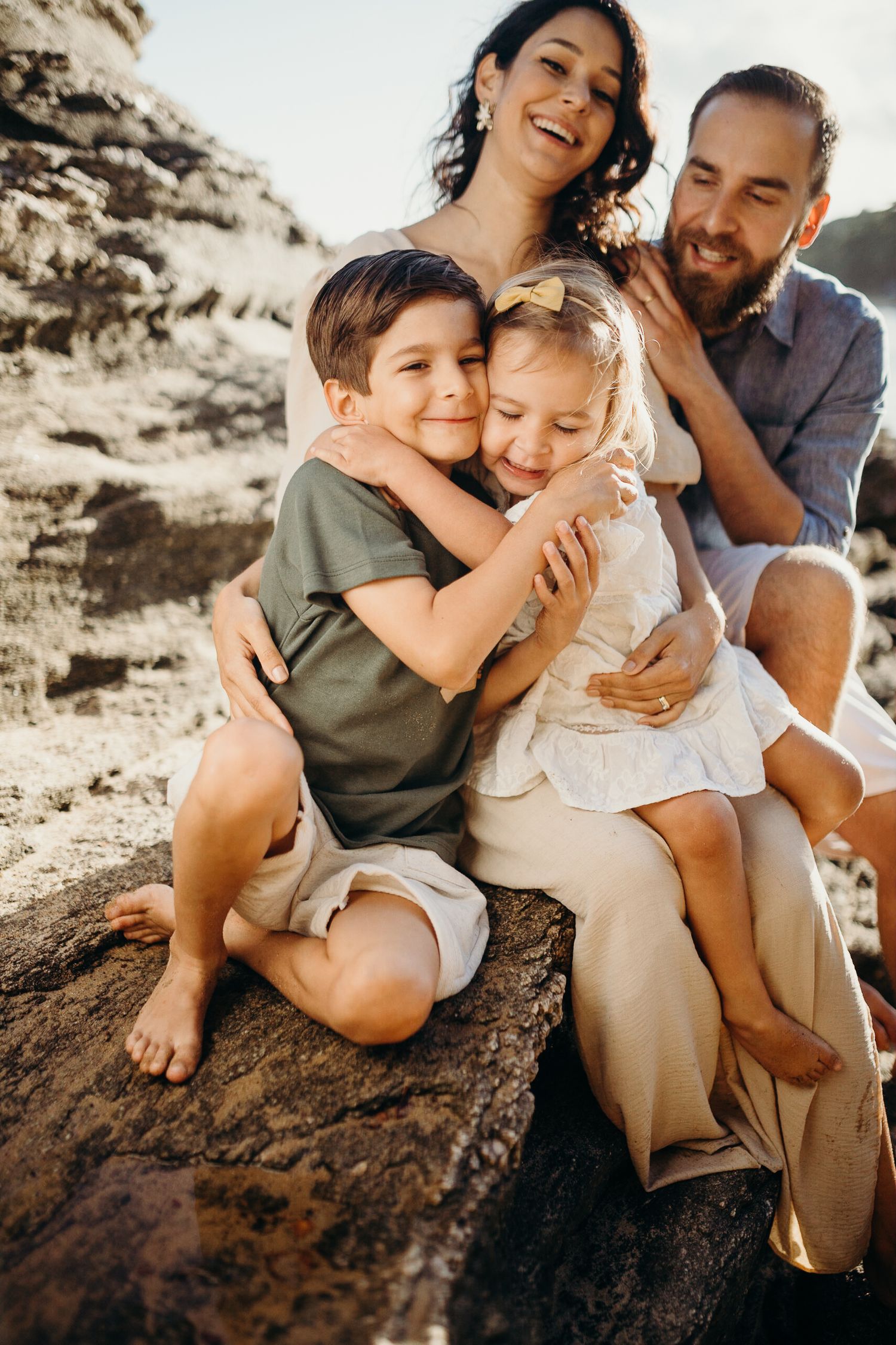 A joyful family sharing laughs and embraces while sitting on coastal rocks.