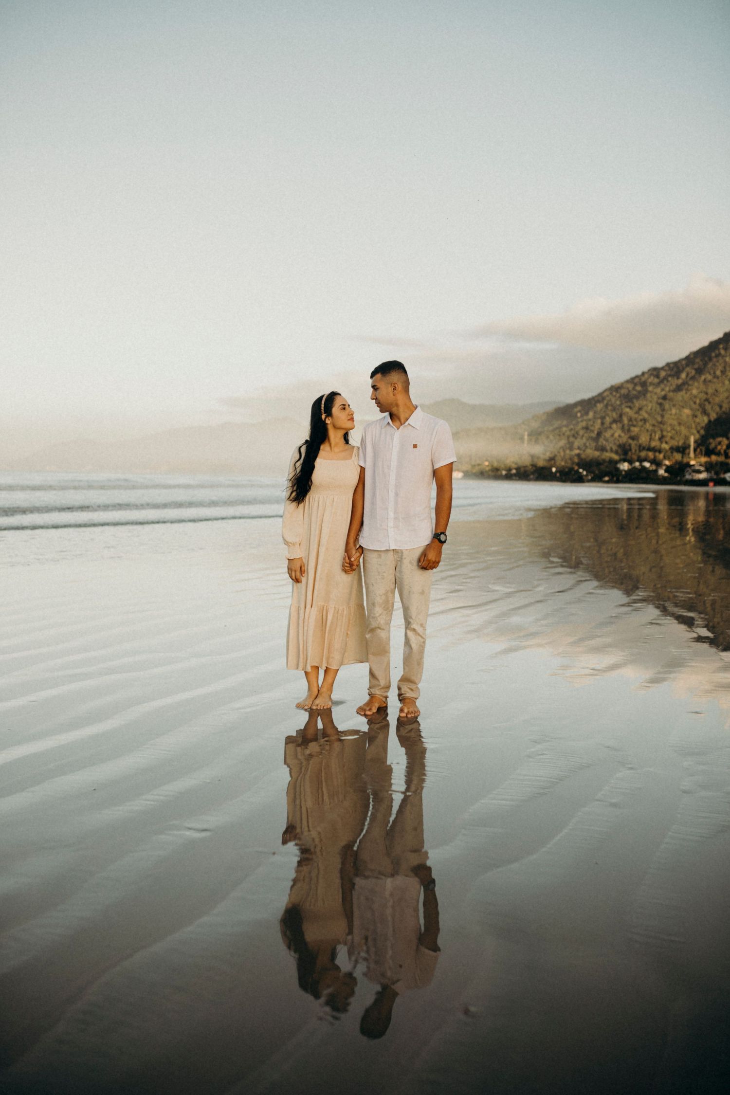 Couple standing on wet sand beach with perfect reflection in water at sunset.