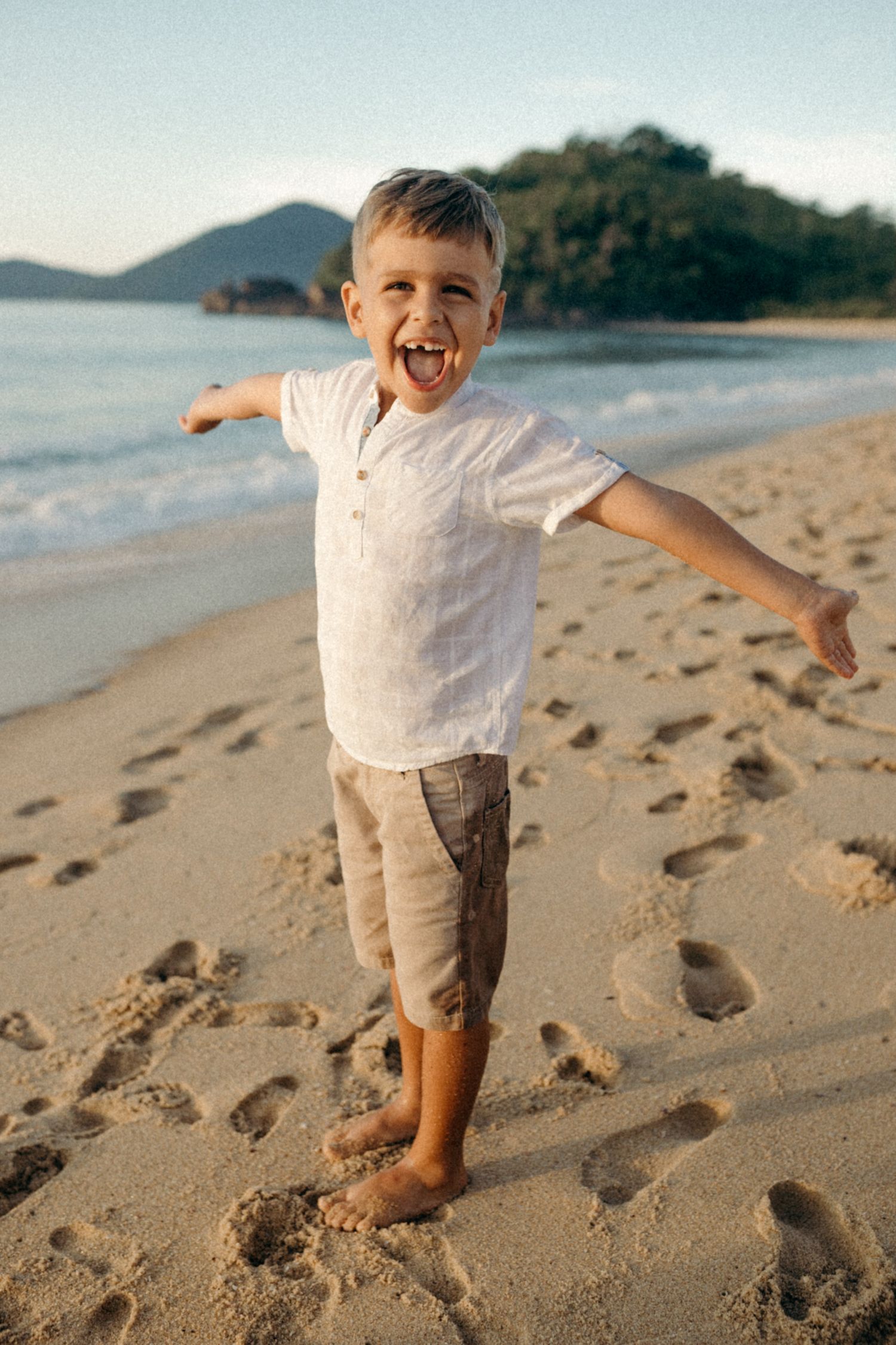 Young child playing joyfully with arms spread wide on sandy beach at sunset.