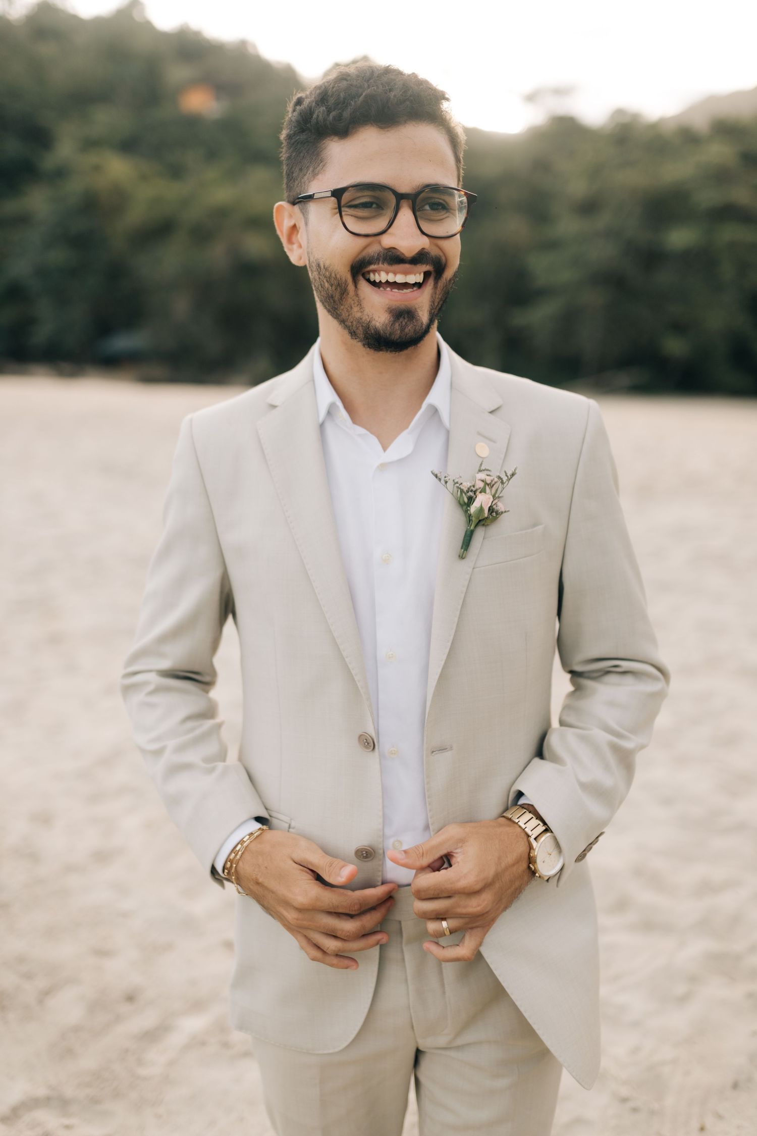 A groom in a light beige suit and glasses adjusts his jacket while standing in an outdoor setting.