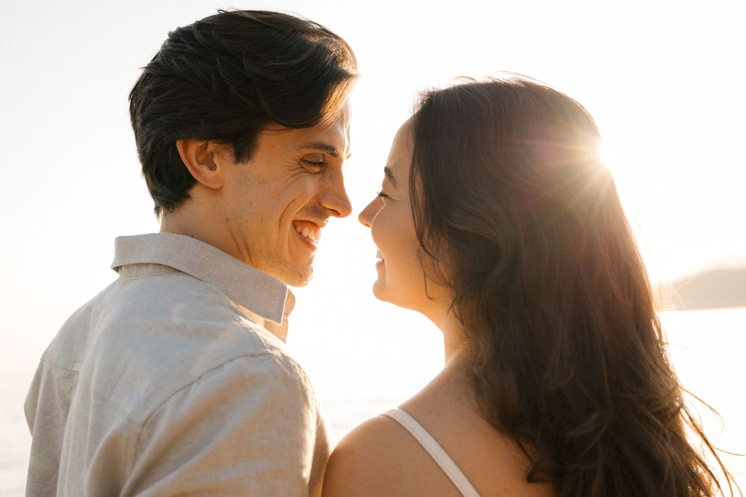 A happy couple stands close together laughing and smiling in the warm sunlight with lens flare behind them.