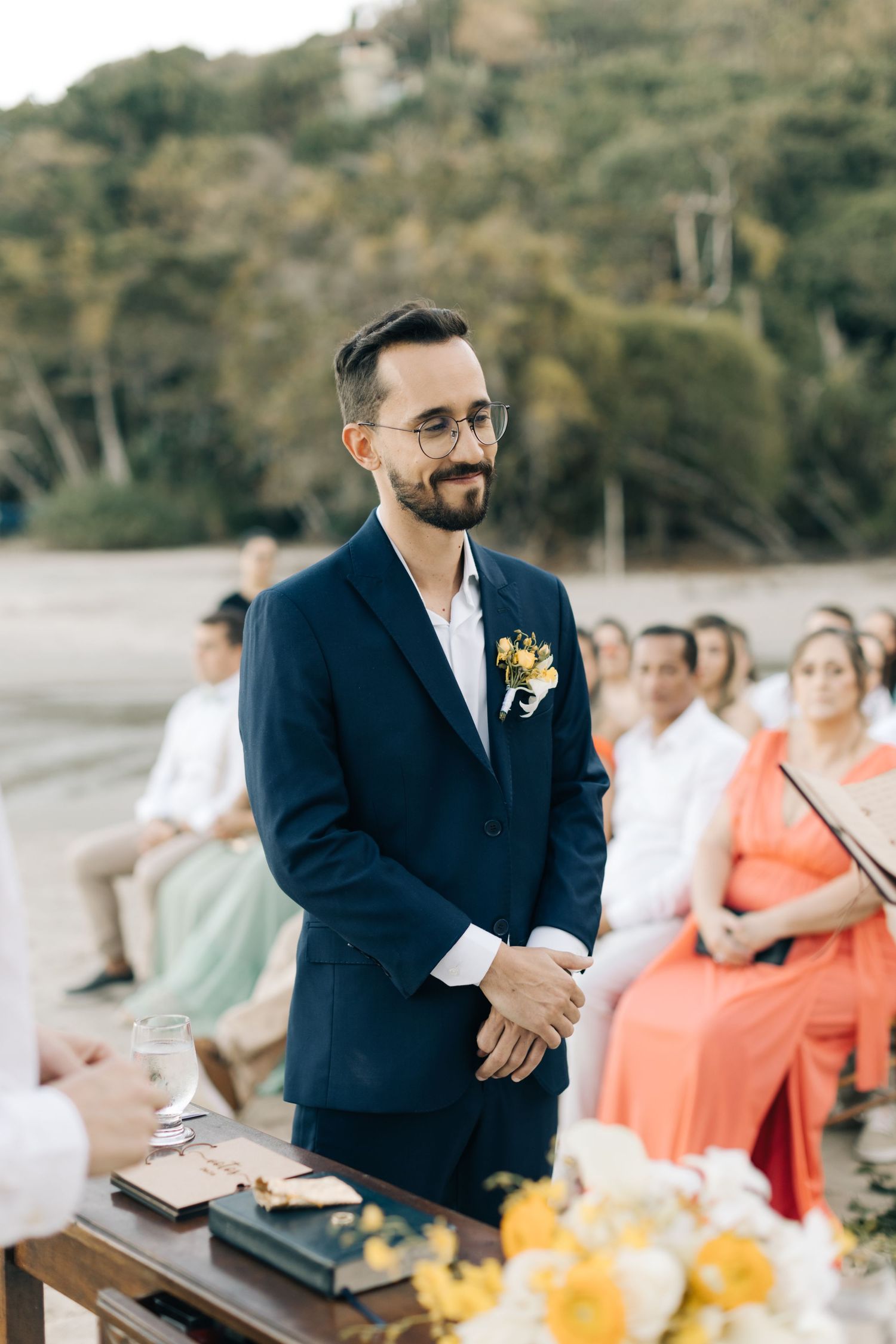 Groom in navy suit stands at wooden podium during beach wedding ceremony.