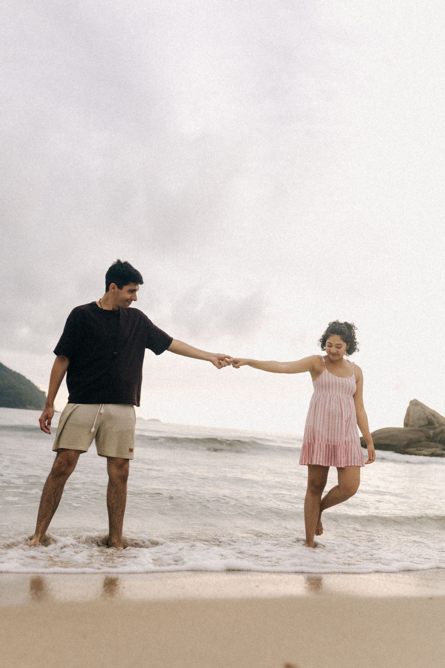 Um casal dá as mãos enquanto está nas ondas do mar em uma praia em um dia nublado.